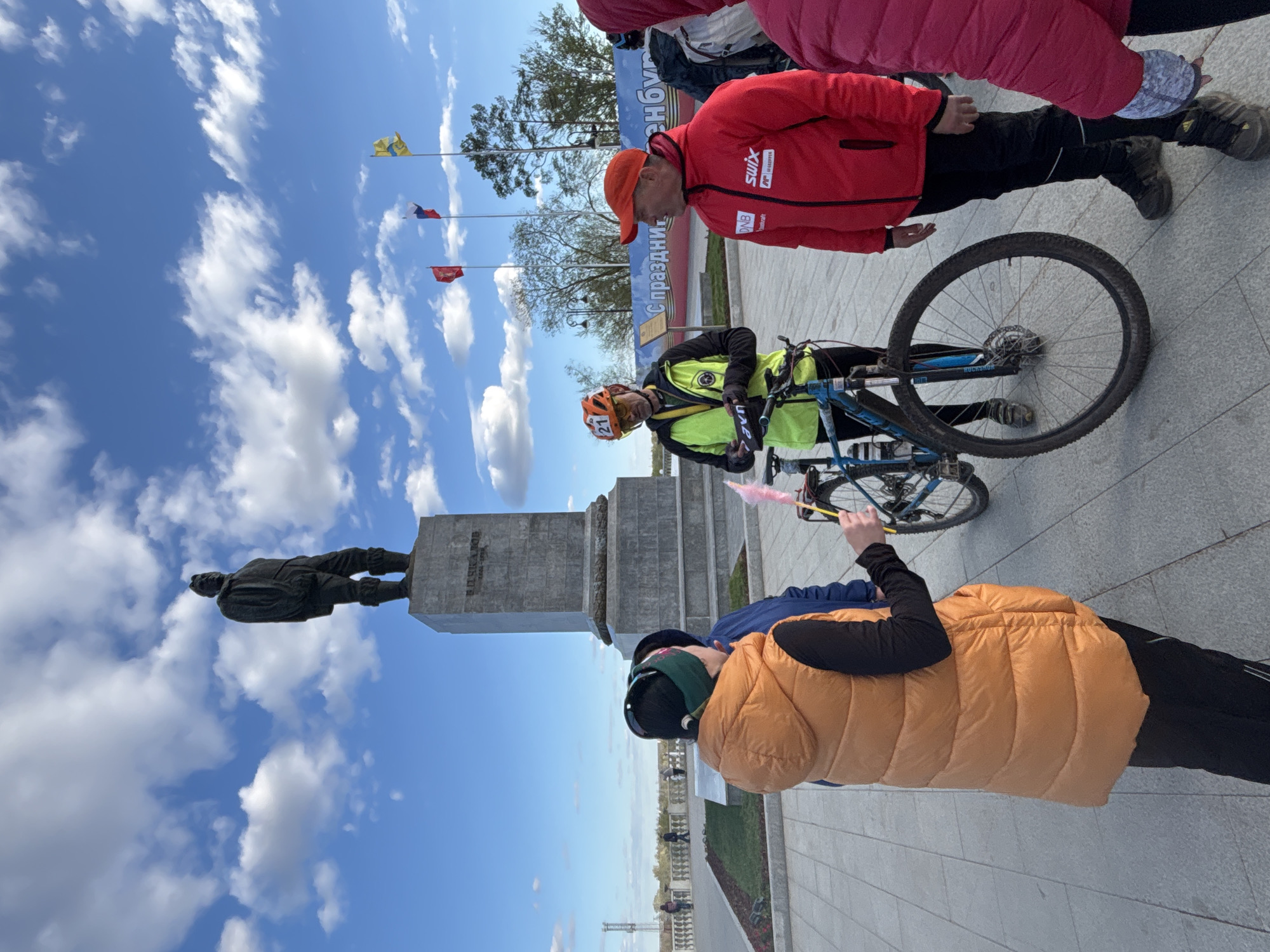 A ground-level, medium, outdoor, natural-lighting photograph depicts a scene in the city of Orenburg, Russia. Three people, including a cyclist, are located near a statue of V.I. Lenin on the embankment of the Ural River.
The cyclist, wearing a yellow and black cycling jersey and an orange helmet with the number "21," stands beside a blue mountain bike. A person in a red jacket and orange cap is standing near the cyclist. Another person, dressed in an orange puffy vest, is holding pink cotton candy.
The statue of Lenin is on a tall, rectangular, concrete plinth. Flags are waving in the background.
The sky is blue with scattered white clouds. The ground is paved.