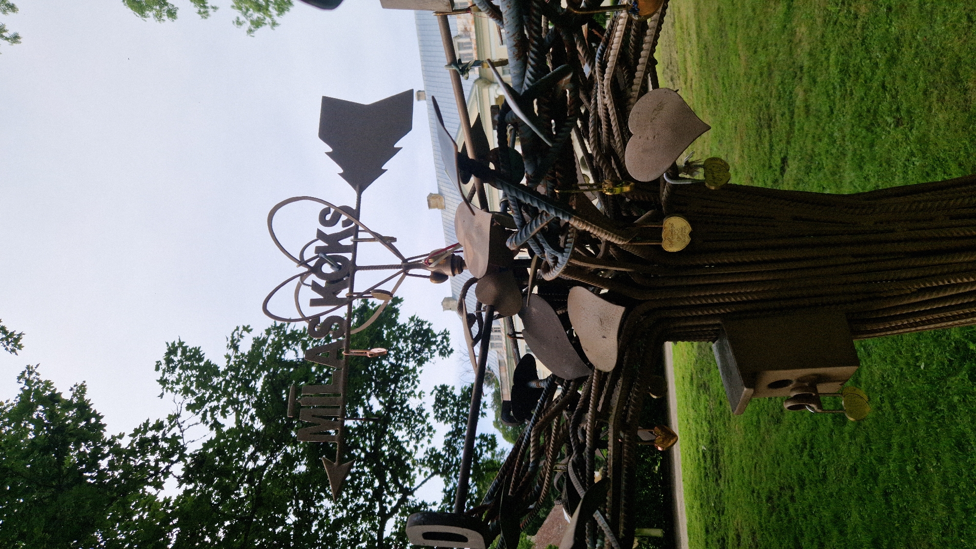 The image depicts a sculpture made of metal rods and rebar, shaped like a tree. The sculpture is adorned with numerous heart-shaped locks and decorative elements, including a birdhouse. At the top of the sculpture, a metal sign reads "MĪLAS KOKS" (Tree of Love) with an arrow and a heart shape. The sculpture is situated in an outdoor setting with green grass and trees in the background. A building with a light-colored facade and a gray roof is visible behind the sculpture, suggesting an urban or park environment. The sky is overcast. This sculpture is located in the city of Kazdanga, Latvia.