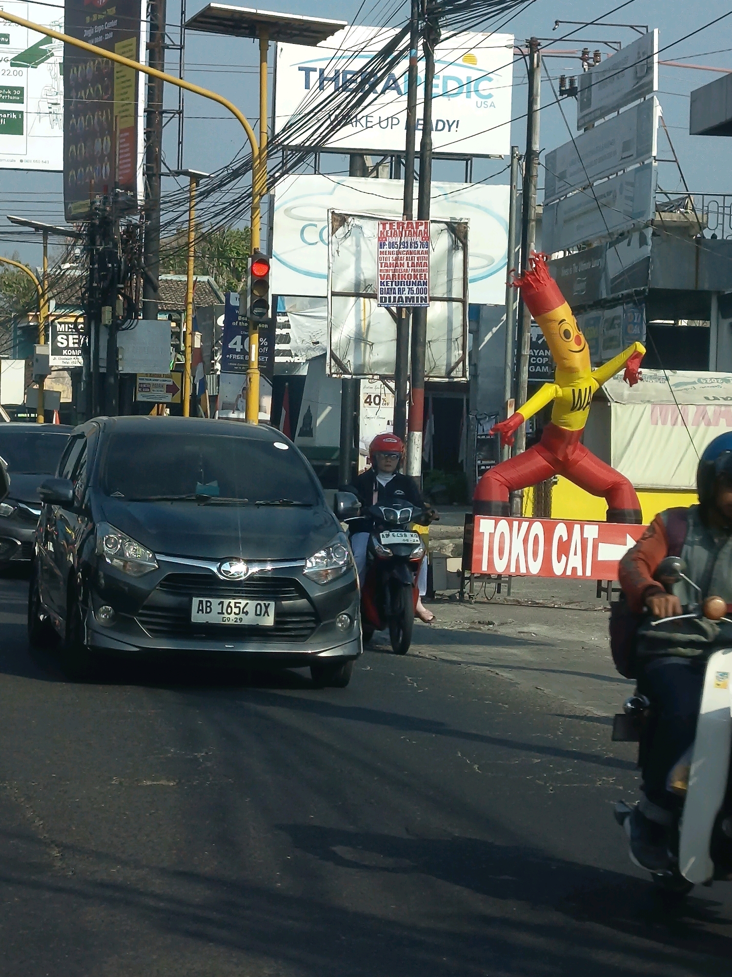 The image captures a street scene in Sidoarum, Indonesia, focusing on a traffic intersection with a red traffic light. A dark grey sedan, bearing the license plate AB 1654 OX, is stopped in the foreground, facing forward. To the right of the sedan, a person on a red and black motorcycle is also stopped. Further down the street, a large inflatable tube man in yellow and red attire stands prominently, advertising "TOKO CAT" (Paint Store) with an arrow pointing to the right. Behind the inflatable man, a white building with "TOKO CAT" signage is visible. Various billboards and signs are present throughout the scene, including one for "THE MEDIC USA" and another advertising a discount of "40% off." The environment appears to be a busy urban area with overhead wires and a clear sky.