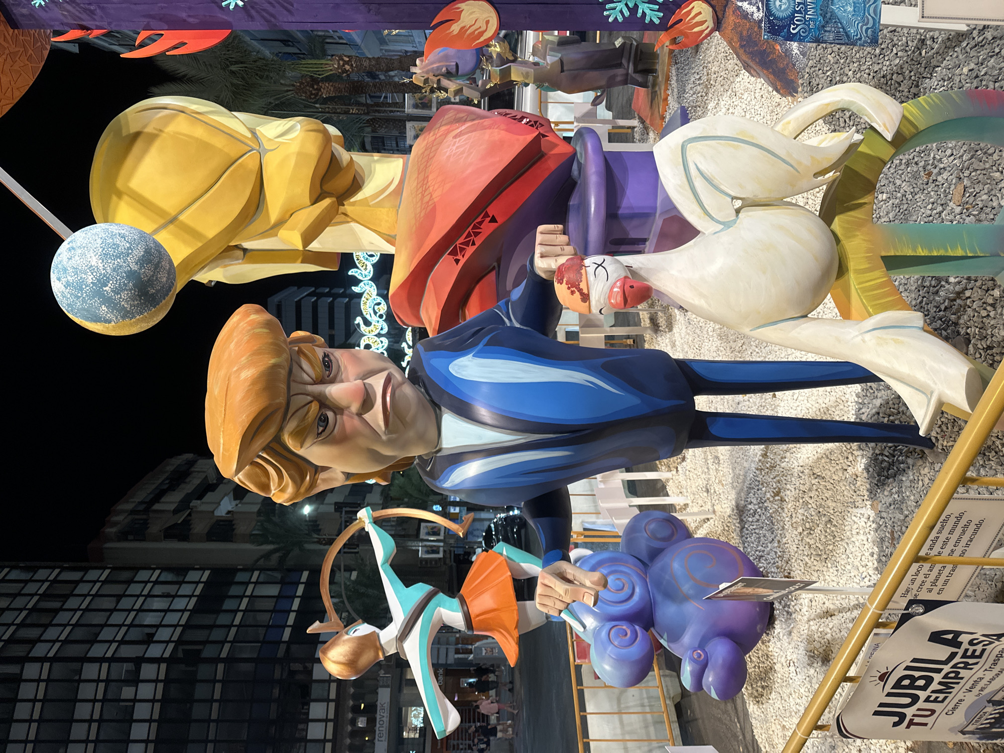 The image shows a street scene at night in Alicante, Spain, featuring large, colorful sculptures. The central sculpture is a caricature of Donald Trump, depicted with an orange wig and an angry expression, wearing a blue suit. To his left is a sculpture of a woman dressed as an archer, holding a bow. To the right of the Trump figure is a large, abstract yellow sculpture topped with a blue and yellow orb. In the foreground, a white goose-like sculpture with a red beak is perched on a colorful base. Several palm trees and buildings are visible in the background. The ground is covered with gravel. There are barriers and signs, including one in Spanish that says "JUBILA TU EMPRESA". The scene appears to be part of a festival or public art installation.