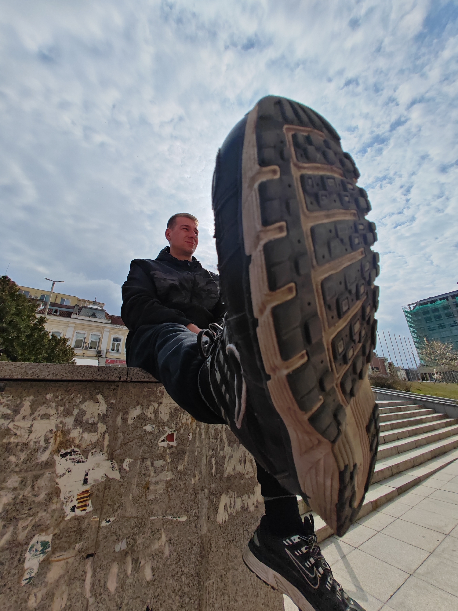 A low-angle photograph captures a single male subject seated on a concrete retaining wall in Shumen, Bulgaria. The subject, positioned to the left of the frame, is a young adult with short, light hair, wearing a black hooded jacket with a camouflage pattern and dark trousers. He is looking towards the right with a neutral expression. His left leg is extended towards the camera, prominently displaying the dark and beige textured sole of a sports shoe in the upper right quadrant. The wall upon which he sits shows remnants of torn paper and adhesive. In the background to the left, several older, light-colored multi-story buildings with traditional roofs are visible. To the right, a wide set of concrete stairs ascends, and a modern multi-story building under construction, wrapped in green scaffolding, stands further back. A series of thin, dark vertical poles are also present in the mid-ground. The sky is partially cloudy with scattered white clouds. The setting is identifiable as the urban area around Ploshtad Bulgaria in Shumen, with the stairs leading towards the "1300 Years Bulgaria" Monument complex.