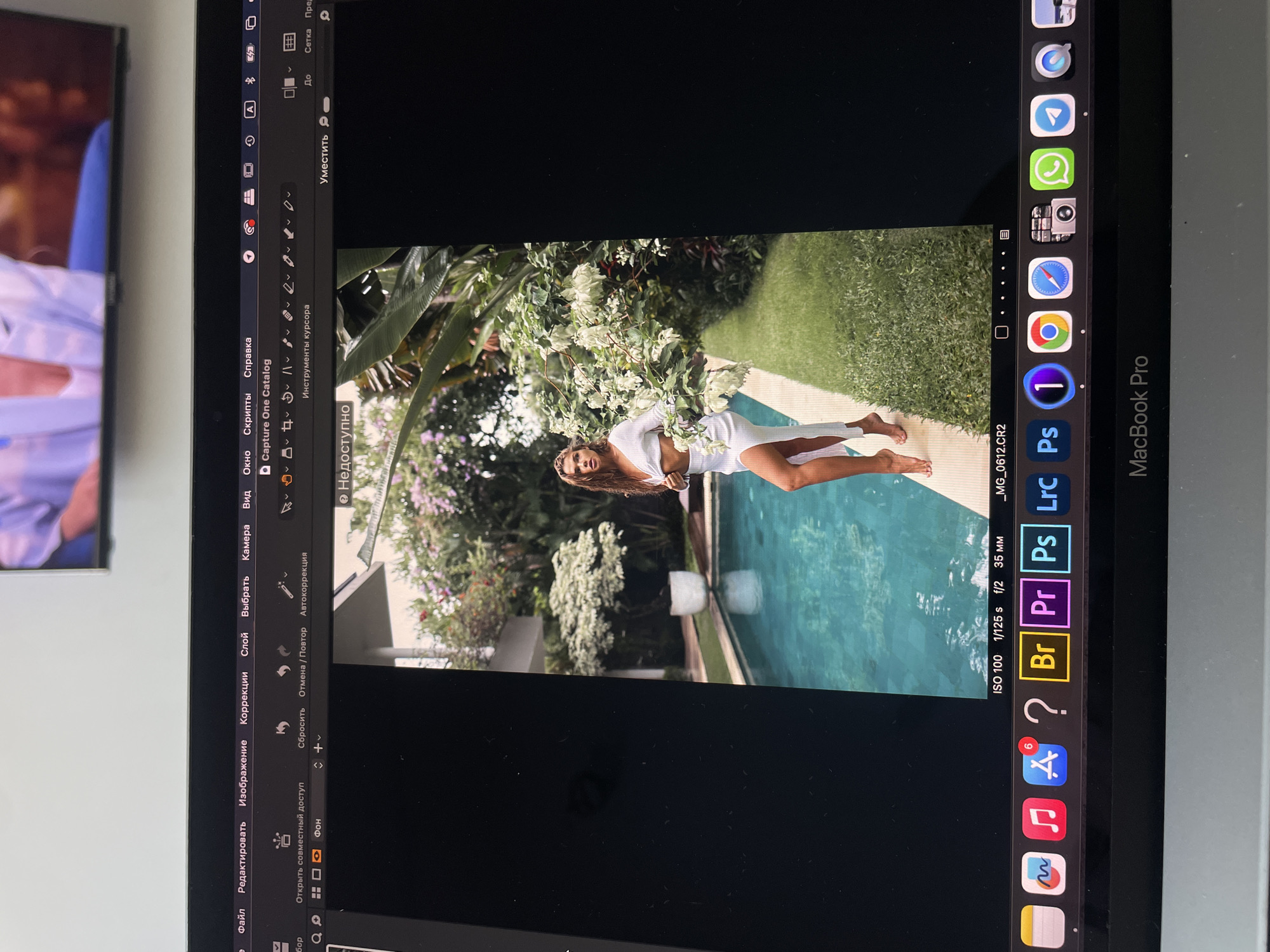 The image shows a MacBook Pro screen displaying an image being edited in photo editing software. The photo features a woman in a white dress posing by a pool surrounded by greenery. The right side of the screen displays the macOS dock with icons for applications such as Adobe Photoshop (Ps), Capture One (1), Safari, Google Chrome, WhatsApp, Telegram, and other standard macOS applications. On the left side of the screen, the photo editing software's menu bar is visible, with options like "File," "Edit," "Image," "Corrections," "Layer," "Select," "Camera," and "View," written in Russian. Below the menu bar, there are additional tool options. The photo editing software is Capture One catalog. The image also displays the camera settings used to take the photograph such as ISO 100, 1/125 s, and f/2 at 35mm. There is a television in the background. The image location is not specified in the image.