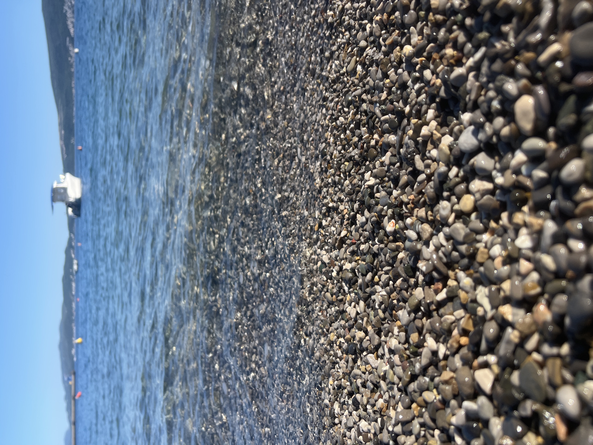 The image shows a close-up view of a pebble beach in Bijela, Montenegro, with the Adriatic Sea lapping at the shore. The pebbles are mostly gray and tan, wet from the receding waves. In the distance, a white boat is anchored in the clear, blue water. The coastline is visible in the background under a cloudless sky. Small orange and yellow buoys are sparsely floating to demarcate an area for the swim.