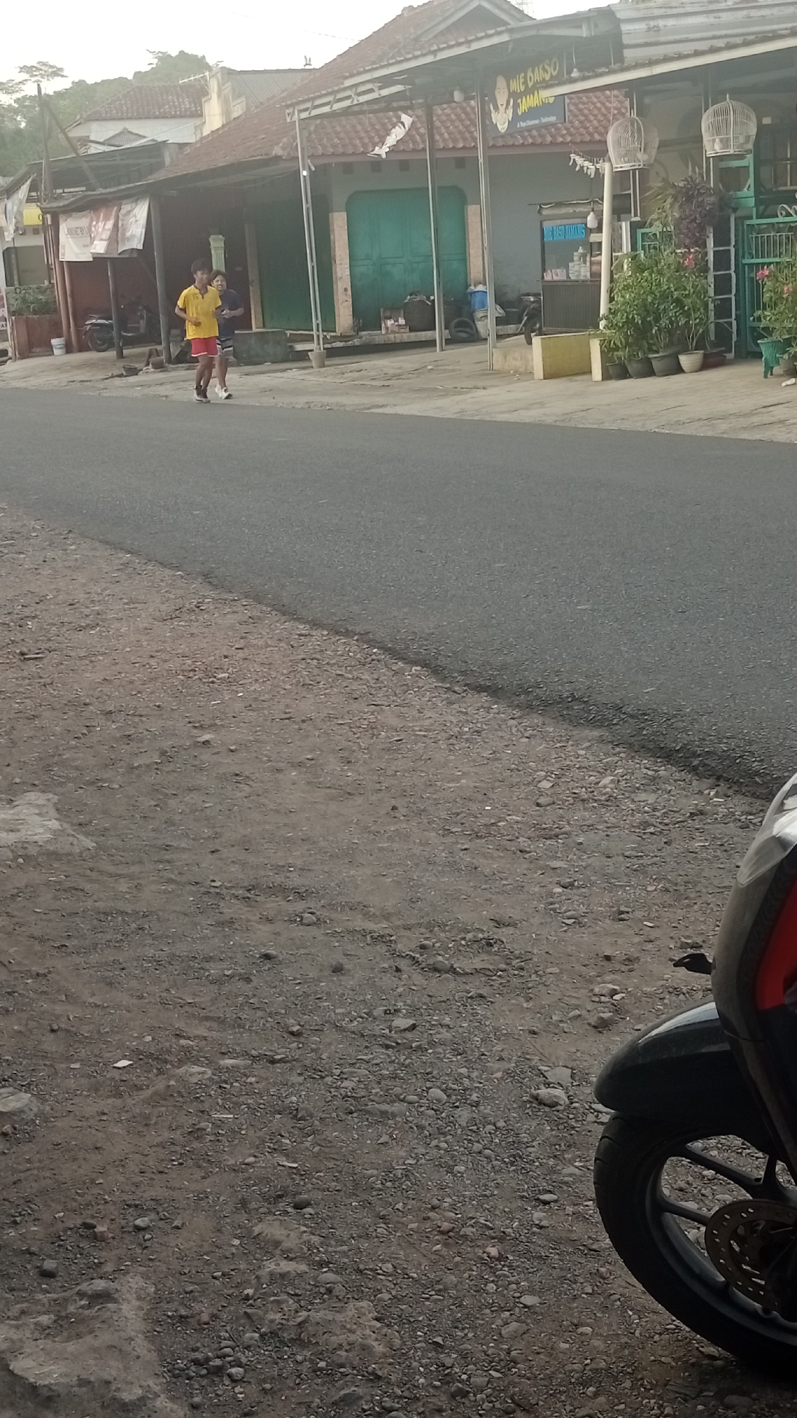 In Pakemitan, Indonesia, two young men are jogging on the side of a paved road in front of a row of buildings. The man in the foreground, on the left, wears a yellow t-shirt and red shorts, while the man behind him wears a dark blue shirt and grey shorts. The buildings to their right appear to be shops or residences with open fronts. One building has a sign that reads "MIE DARSO JAMAN NOW." Various potted plants and bird cages are visible outside the establishments. A motorcycle is parked in the foreground on the right side of the image, partially obscuring the dirt and gravel shoulder of the road. The overall scene depicts an everyday activity in a semi-urban environment.