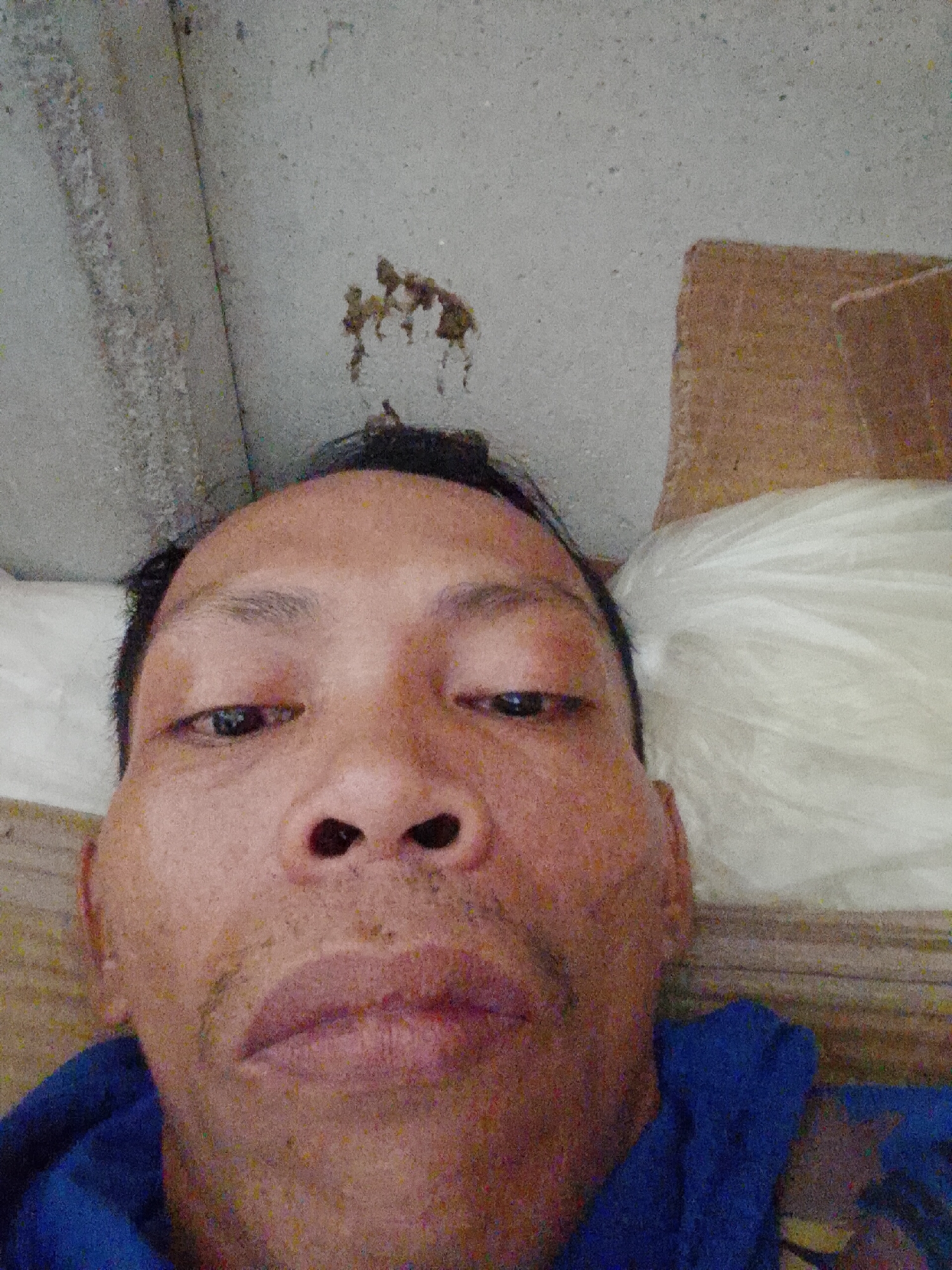 A close-up selfie of a man lying down, possibly on a bed, is captured indoors. The man has short black hair and a brown complexion. He is wearing a blue shirt. The background includes a white pillow, a brown object that could be part of the bed frame, and a ceiling with a brown stain. The image appears to be taken from a low angle, looking up at the man's face. The location is Bukit Ketok, Indonesia.