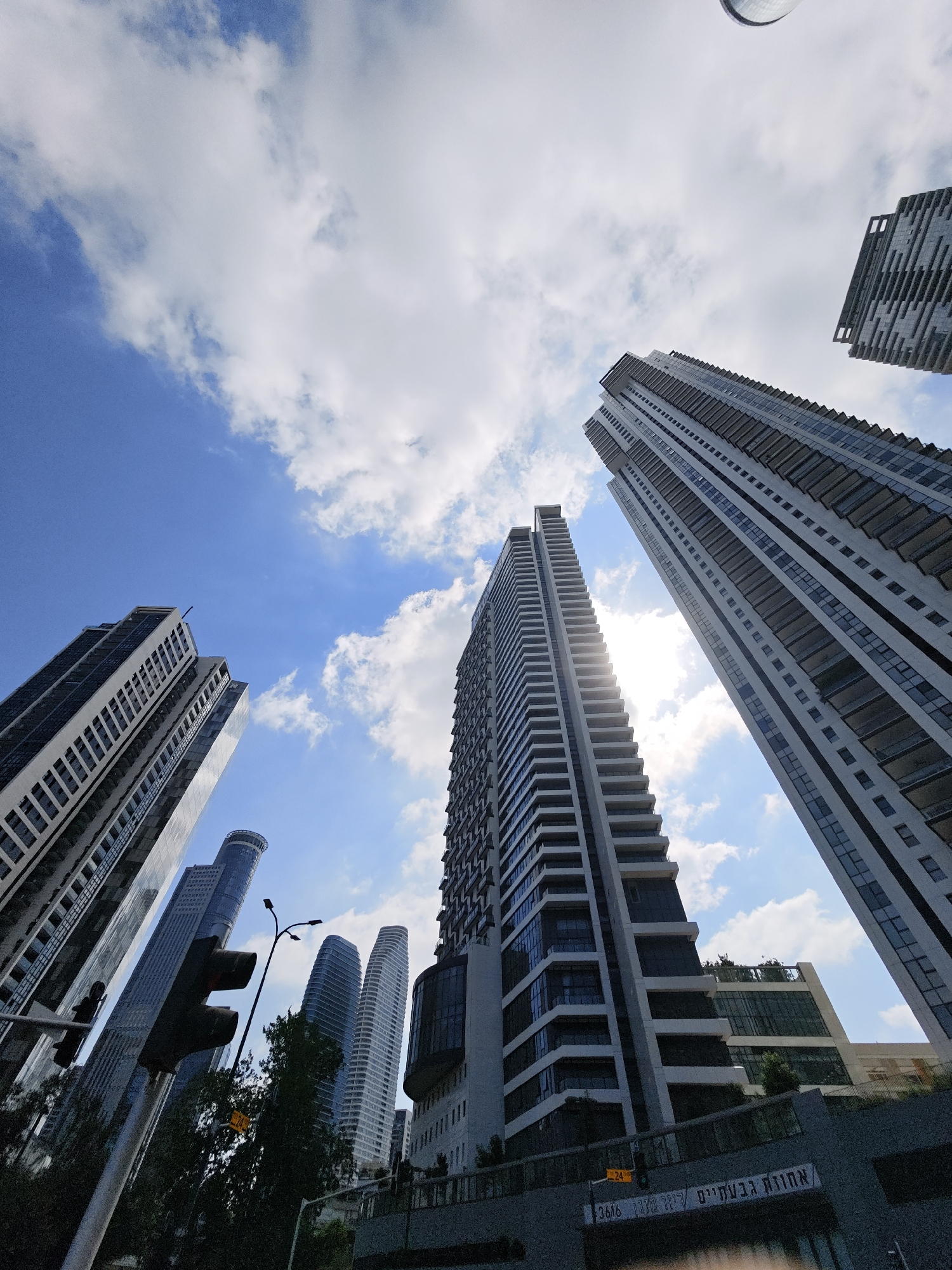 The image captures a low-angle view of a city skyline, dominated by modern high-rise buildings under a bright, partly cloudy blue sky. The architecture is characterized by sleek lines, large windows, and numerous balconies. Several prominent skyscrapers are visible, some appearing to be residential or office towers with repeating patterns of windows and balconies.
In the foreground, on the left side, a traffic light pole with multiple signal heads is prominently featured, along with a streetlight. Some greenery, likely trees, is visible below the traffic light. The base of some buildings shows commercial or public spaces, with visible signage.
One of the buildings in the background has text written on it in Hebrew. Based on the provided context, this scene is located in Givatayim, Israel. The overall impression is one of urban development and contemporary architecture, set against a clear sky. There are no people visible in the image.