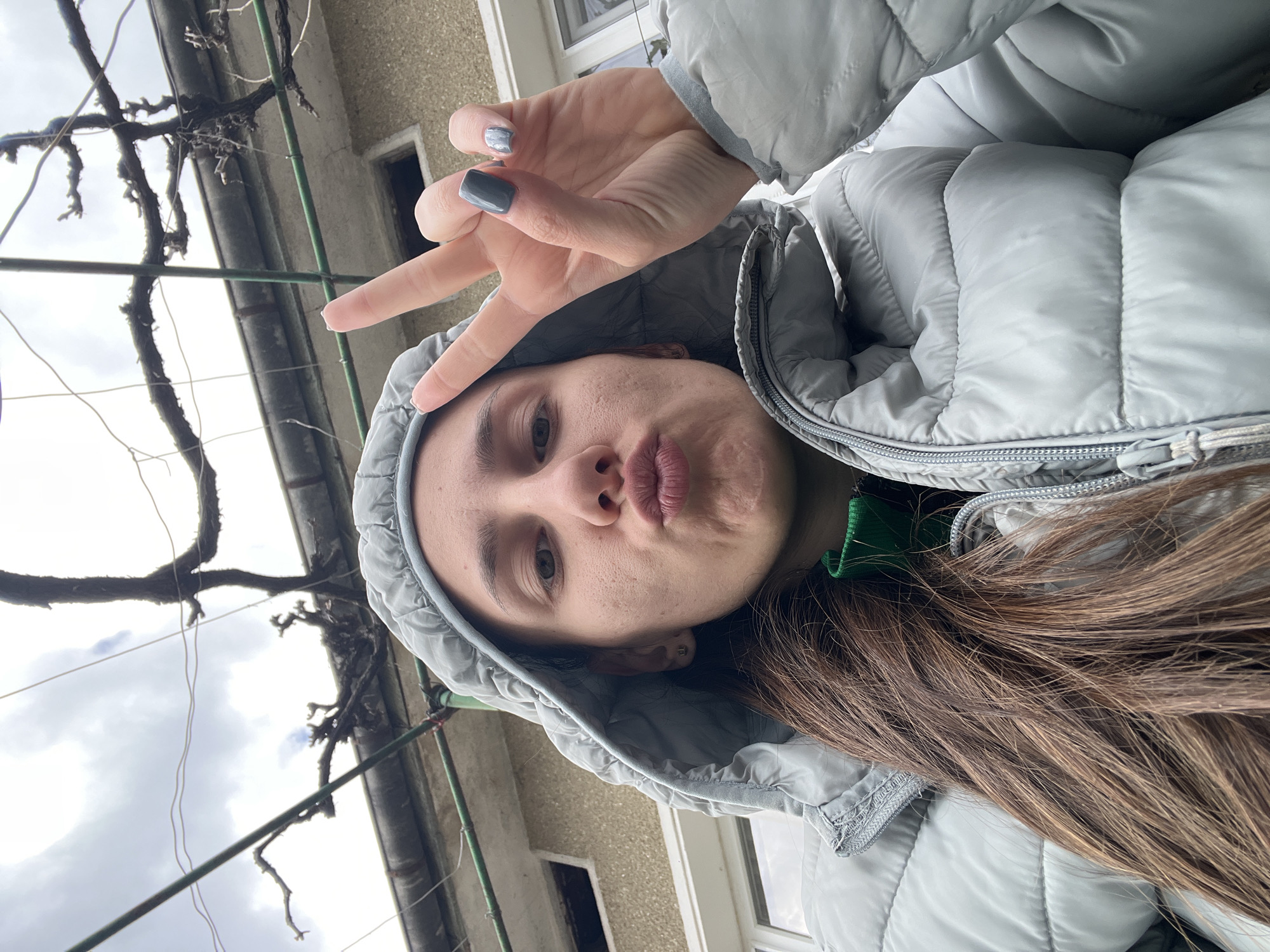 A woman is outdoors, wearing a grey puffer jacket with the hood up. She is making a peace sign with her left hand and puckering her lips. She has brown hair. In the background, there is a building with a window and a bare vine growing along the structure. Based on the location provided, this scene is in Hrishteni, Bulgaria.