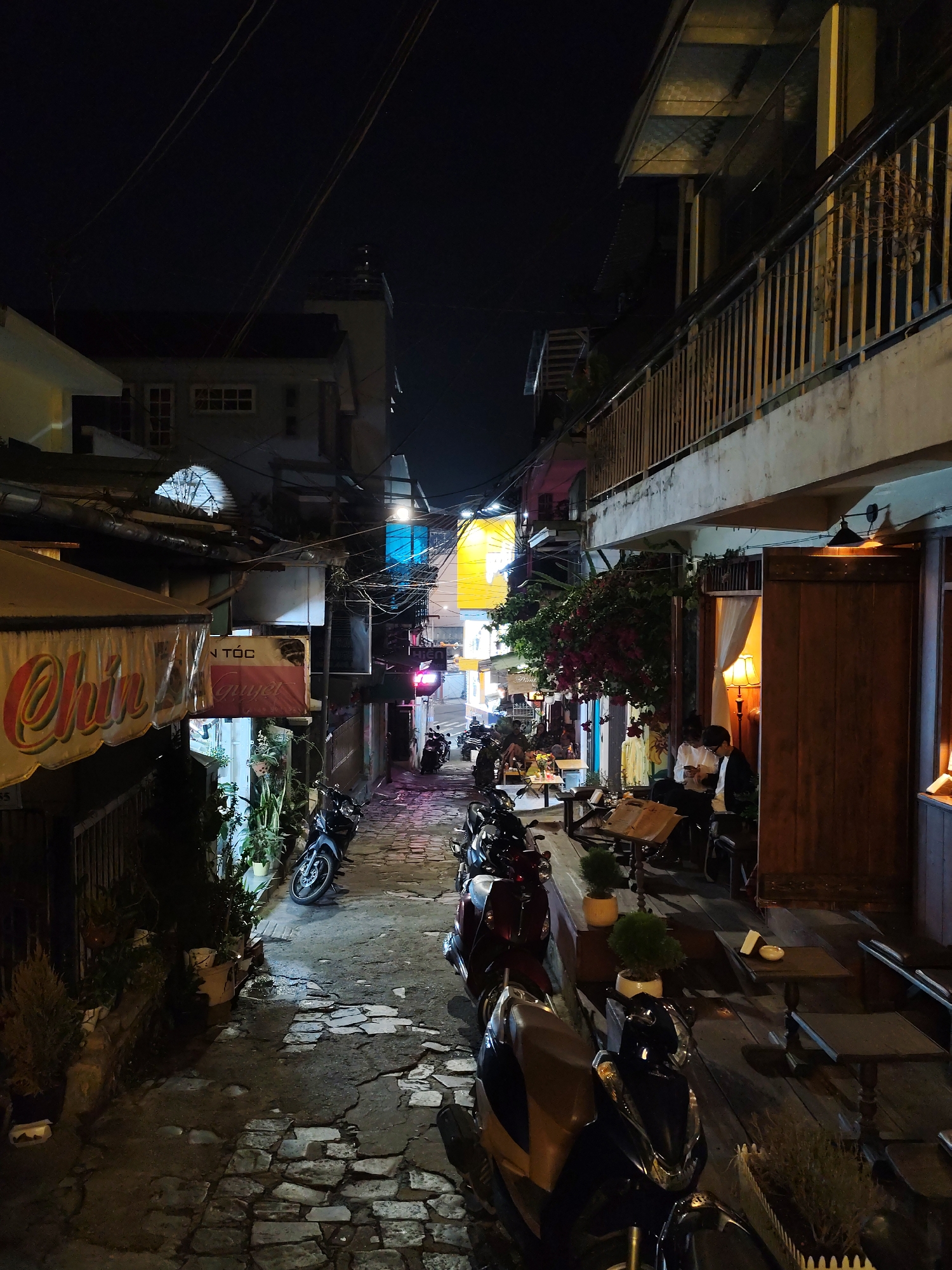 The image depicts a narrow alley in Da Lat, Vietnam, at night. The alley is paved with cracked stone and lined with buildings on either side. Several motorbikes are parked along the alley. On the right side of the alley, there is a building with a balcony. A couple sits at a table outside an establishment with wooden doors, illuminated by a table lamp inside. On the left side of the alley, there are signs for businesses, including "Chin" and "An Toc." Electrical wires crisscross overhead. Further down the alley, there are additional tables and motorbikes, with more buildings visible in the distance, partially lit by colorful lights. The sky above is dark.