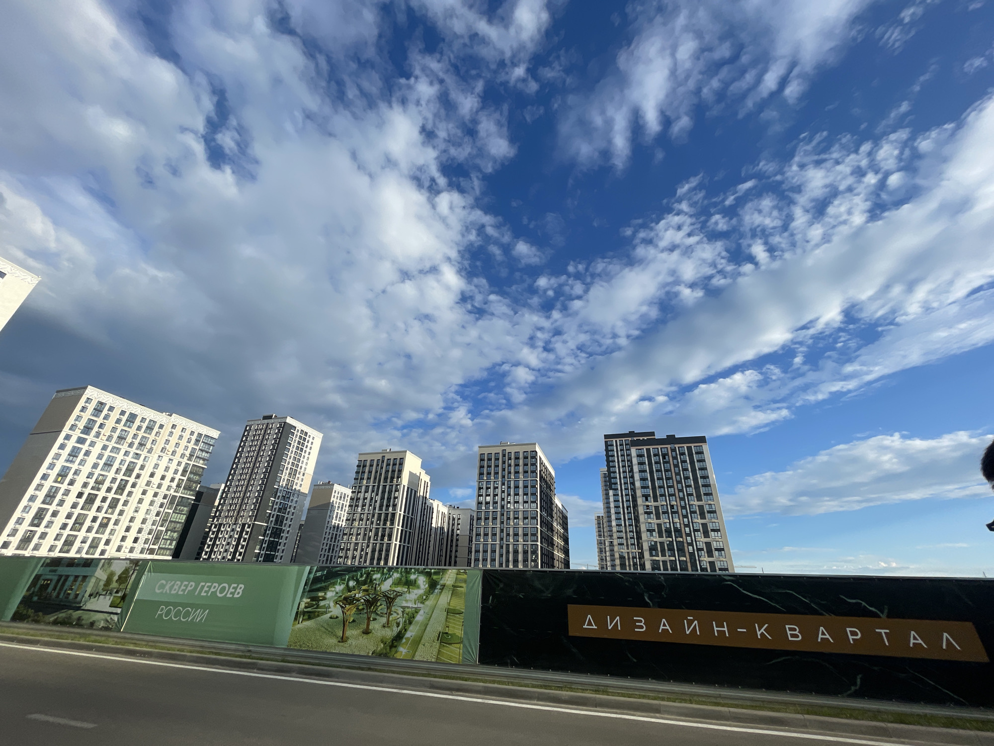 The image shows a construction site in Stavropol, Russia, with several modern high-rise apartment buildings under construction. The buildings are tall and feature geometric designs with many windows. A large banner runs along the front of the site, displaying renderings of the finished development and text in Russian. One section of the banner reads "СКВЕР ГЕРОЕВ РОССИИ" which translates to "Square of Heroes of Russia." Another section of the banner reads "ДИЗАЙН-КВАРТАЛ" which translates to "Design Quarter." The sky is a bright blue with scattered white clouds. The foreground shows a section of a road, likely an adjacent highway or street, with a curb and a grassy median. The perspective is from ground level, looking up at the buildings and sky.