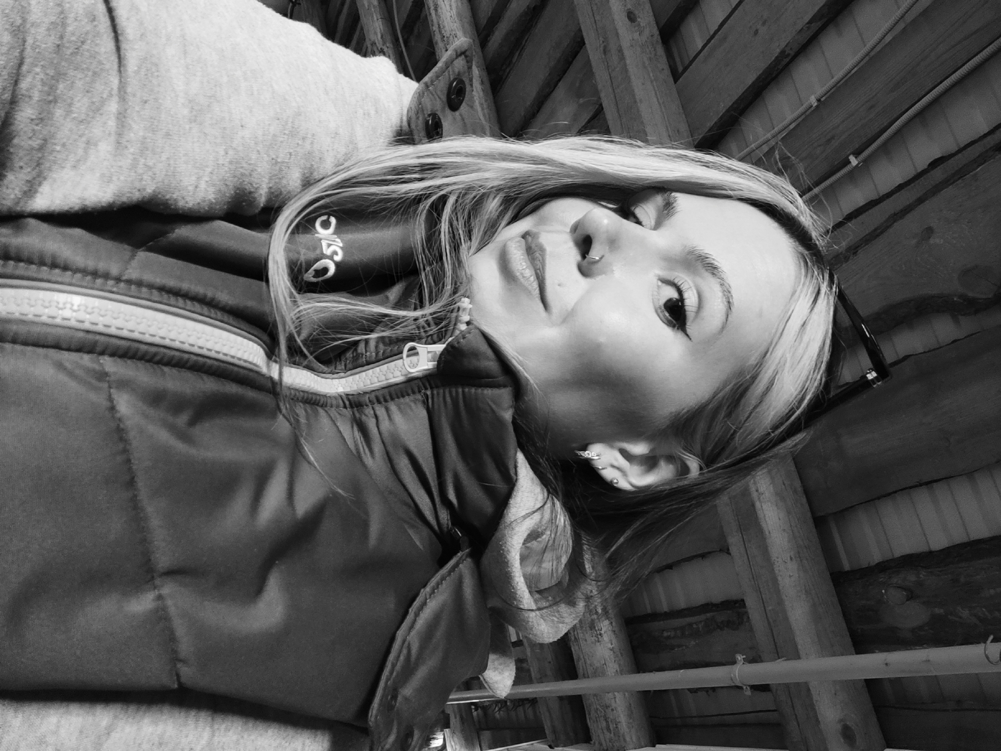 A black and white, close-up portrait shows a young woman looking upwards towards the camera from an angled perspective. She appears to be lying down or reclining against a surface.
**Subjects:** The primary subject is a woman with light-colored hair, possibly blonde, which frames her face. She has a subtle nose piercing, a small hoop in her left nostril. Her lips are full, and her eye makeup, particularly eyeliner, is visible. She wears a light-colored, possibly grey, long-sleeved top, layered under a dark, padded jacket or vest. A prominent zipper is visible on the jacket. On the dark garment, near her left shoulder/neck area, there is text that appears to read "ОЅІС" or "OSIC" or "OSC". A pair of sunglasses is perched on her head or behind her right ear.
**Setting:** The background features a rustic wooden structure, consisting of planks, beams, and possibly corrugated material, suggesting an attic, a wooden ceiling, or an unfinished interior space. Several cables or ropes are visible running across the wooden elements. The location context provided is Ольховка, Russia.
**Activity:** The woman is posing for a photograph, looking directly at the camera with a relaxed and soft expression.
**Notable details:** The image is monochromatic. Her gaze is gentle, and her expression conveys a sense of calm. The wooden background creates a natural, unrefined ambiance. The time of day and weather are not discernible due to the indoor setting and black and white format.