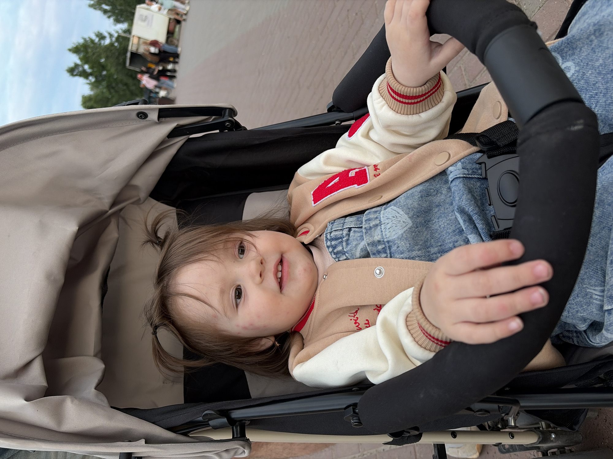 A young child sits in a stroller, looking to the right with a slight smile. The child has brown hair styled in two small ponytails, rosy cheeks with some red spots, and is wearing a light-colored jacket with red trim and an embroidered red "A" on the chest, over denim overalls. The stroller is beige and black. In the background, an outdoor scene is visible with trees, a paved walkway, and several people standing near a white truck or trailer. The sky is overcast. This image was taken in the city of Chaikovsky, Russia.