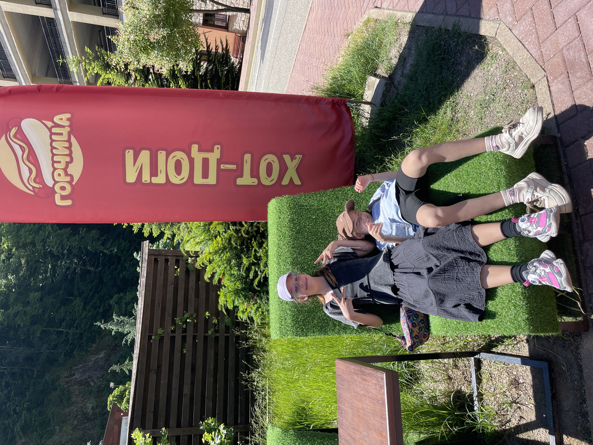 In Estosadok, Russia, two young people are seated on a green, artificial turf bench in front of a large red banner advertising "Hot Dogs" in Russian. The person on the left, wearing a grey shirt, black skirt, glasses, and a purple cap, is making a peace sign with their fingers. The person on the right, wearing a light blue shirt and a brown cap, is holding a bottle and smiling. A small wooden table is to the left of the bench. In the background, a multi-story building with balconies and a wooded mountainside are visible. The ground is paved with brick and gravel.