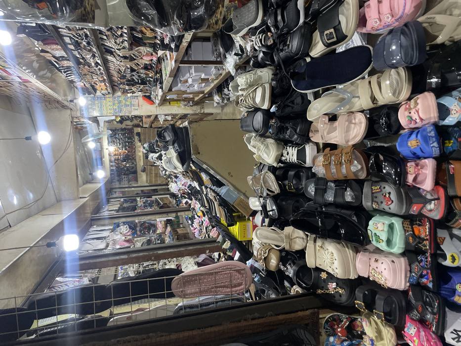 An indoor retail shoe store or market stall is depicted in Nganguk, Indonesia. The scene is brightly illuminated by several bare LED light bulbs hanging from a low, light-colored ceiling. The perspective is slightly elevated, looking down into a densely arranged display of footwear. The foreground and midground are filled with a wide variety of casual shoes, sandals, and slippers in numerous colors, including pink, blue, black, white, brown, and green. Styles include plastic clogs (some with cartoon characters), flip-flops, platform sandals, and children's shoes, arranged closely together on low display surfaces. A single pink-soled slipper or sandal hangs vertically on a metal rack on the left. The background features extensive shelving units lining the walls, packed with additional footwear, primarily women's shoes, sandals, and heels, stacked and arranged in rows. White merchandise boxes are visible on lower shelves. A white ceiling fan is partially visible in the upper right. No individuals are present in the image.