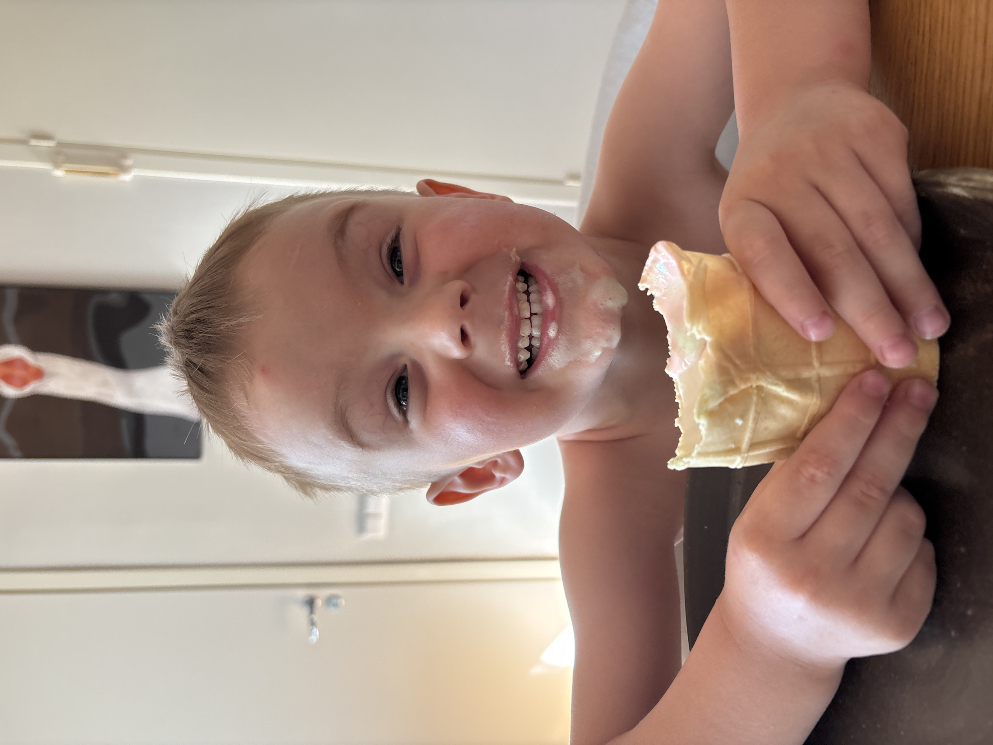 A young, light-skinned child is shown close-up from the chest up, holding an ice cream cone. The child is smiling with visible white teeth and has melted ice cream smeared on their face around the mouth. They are holding the cone in their right hand, with fingers curled around it. The cone appears to be a wafer-type cone.
The background includes a light-colored wall with a piece of dark abstract art hanging on it. There is also a glimpse of a brown surface, possibly a table, at the bottom of the frame.
Based on the metadata, this image was taken in the city of Chaikovsky, Russia. No additional contextual information about the location is apparent from the image.