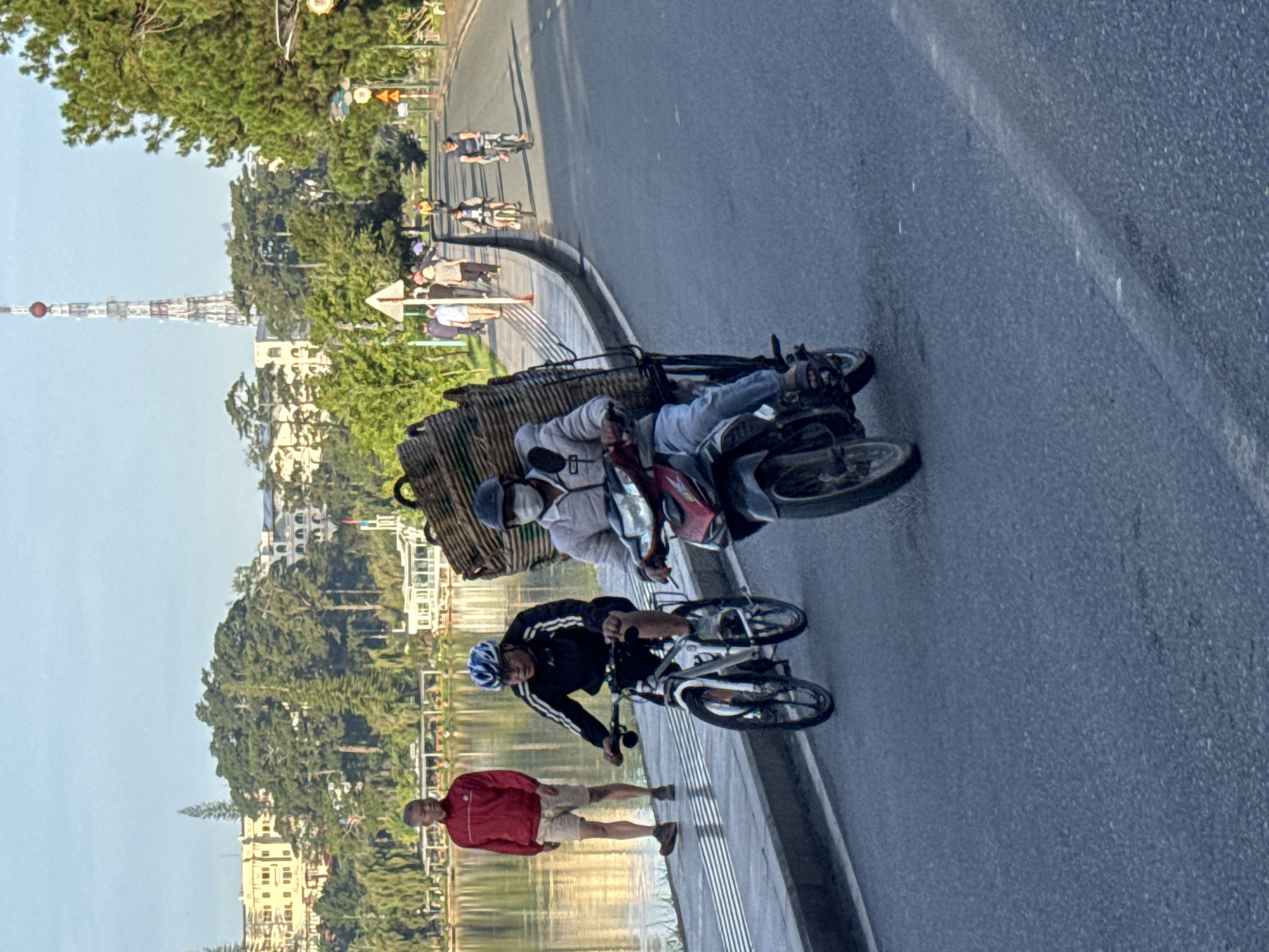 The image depicts a street scene in Da Lat, Vietnam, with a lake and buildings visible in the background. In the foreground, a man wearing a blue helmet and a black jacket is riding a bicycle. He is facing forward and appears to be cycling on a paved road. To his right, another person is riding a scooter and carrying a large woven basket on the back. This person is wearing a helmet and a light-colored jacket. Further back, a man in a red jacket and shorts is standing on a sidewalk next to the lake. Several other people are seen in the distance, some on bicycles and others walking. The environment suggests a temperate climate, with green trees and a body of water. The presence of buildings and infrastructure indicates an urban or suburban setting. The overall scene captures a moment of daily life in Da Lat, with people engaged in various forms of transportation.