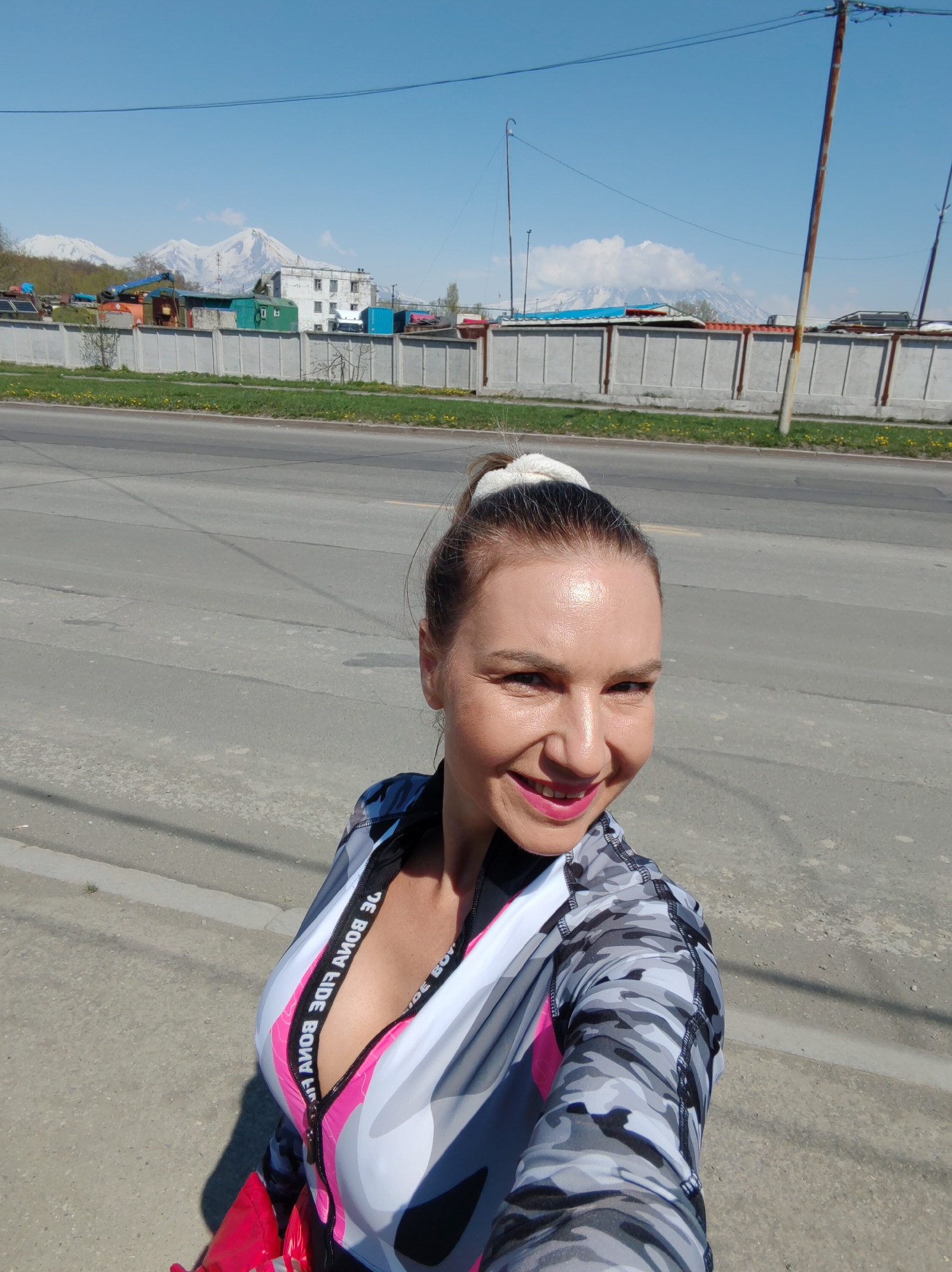 The image captures a selfie of a woman in an outdoor setting, identified as Petropavlovsk-Kamchatsky, Russia, with prominent snow-capped volcanoes in the background.
The woman, positioned in the lower-left to mid-frame, has fair skin, dark hair pulled into a high ponytail secured with a white scrunchie, and is smiling directly at the camera. She has bright pink lipstick and visible facial definition from sunlight. She wears a long-sleeved athletic top featuring a grey, white, and black camouflage pattern, with a prominent pink zipper that exposes a pink undergarment or inner layer. White text, "DE BONA FIDE," is visible and mirrored along the inner lining of the open zipper on both sides.
The immediate background consists of a grey asphalt road in the mid-ground, exhibiting cracks and faint markings. Beyond the road is a narrow strip of green grass interspersed with small yellow flowers. A concrete panel fence, approximately 1.5 meters in height, runs horizontally across the scene. Behind the fence, various structures are visible: an assortment of industrial or storage units, including blue, green, and orange containers or sheds, and a larger, multi-story white building showing signs of wear. Multiple utility poles with overhead wires are present on both the left and right sides of the frame. In the distant background, under a clear blue sky with scattered white clouds, several large, snow-capped volcanoes are prominently featured, characteristic of the Kamchatka Peninsula, specifically the Koryaksky and Avachinsky volcanoes near Petropavlovsk-Kamchatsky. The scene is illuminated by strong sunlight, indicated by shadows cast towards the viewer's right.
