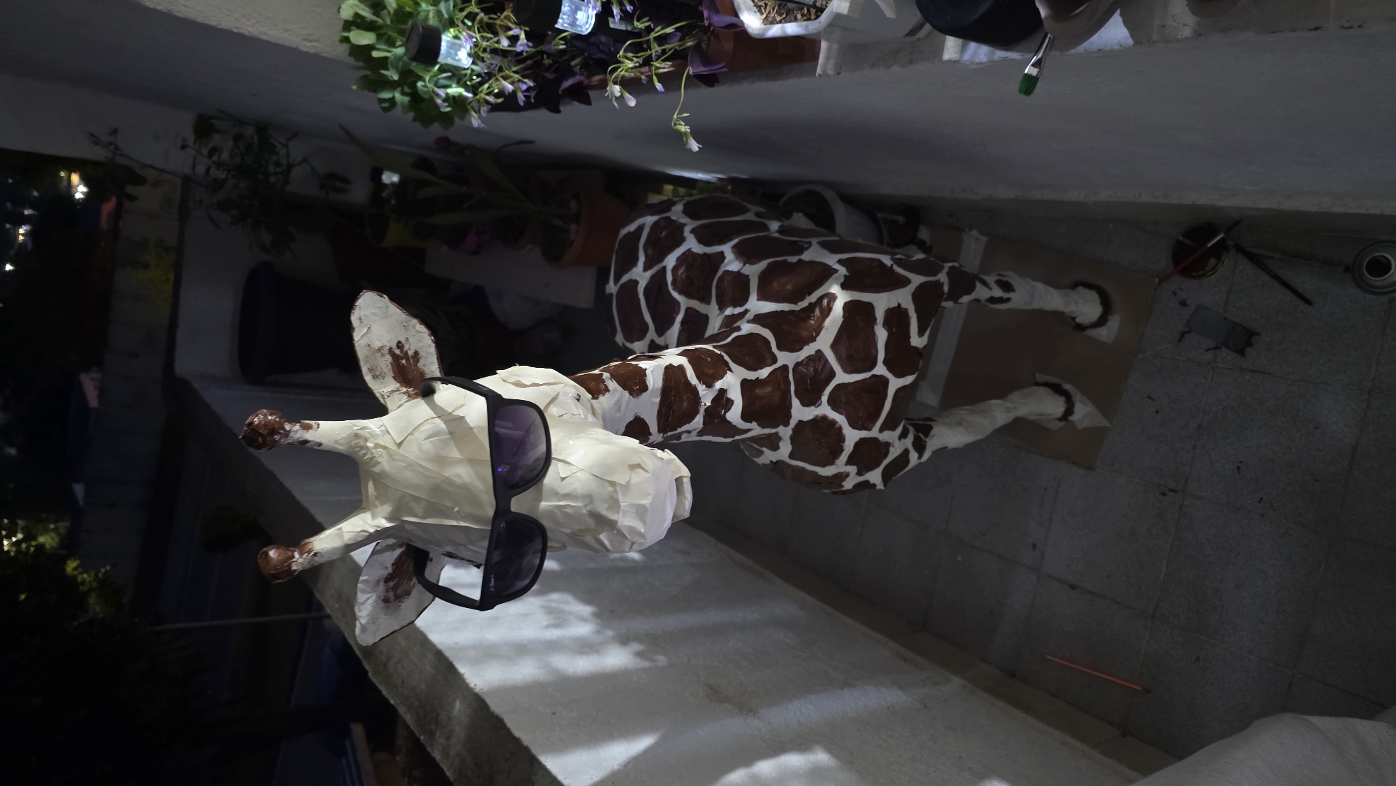 A full-body paper-mache giraffe sculpture wearing sunglasses lies sideways on an outdoor tile floor. The giraffe is mostly white except for brown spots on its body and brown tips on its ossicones. The tiles are light gray. Several potted plants sit on an adjacent ledge. The location appears to be an outdoor area, likely in Blagoevgrad, Bulgaria, given the request.
