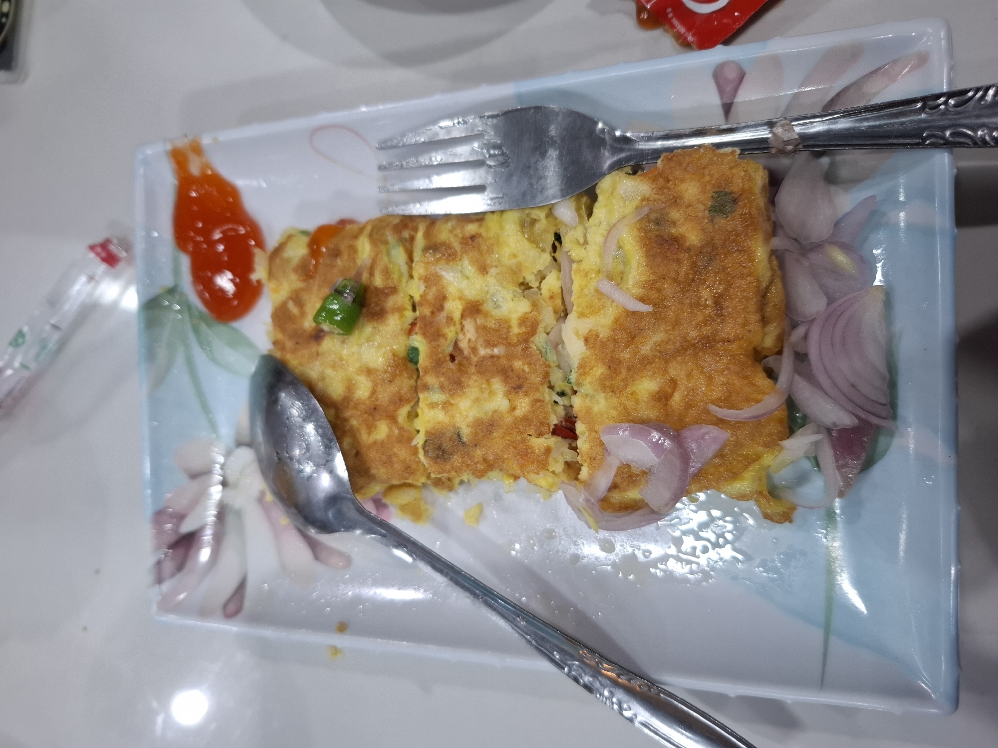 A close-up, top-down shot shows a rectangular, blue and white patterned plate of food. The plate is filled with a sliced omelet, topped with sliced red onions and a dollop of red chili sauce. A fork and spoon are positioned on the right and left sides of the plate, respectively. The food appears to be on a white, reflective surface, possibly a table. The overall scene suggests a meal being served in a casual dining setting in Kampongbaro, Indonesia.