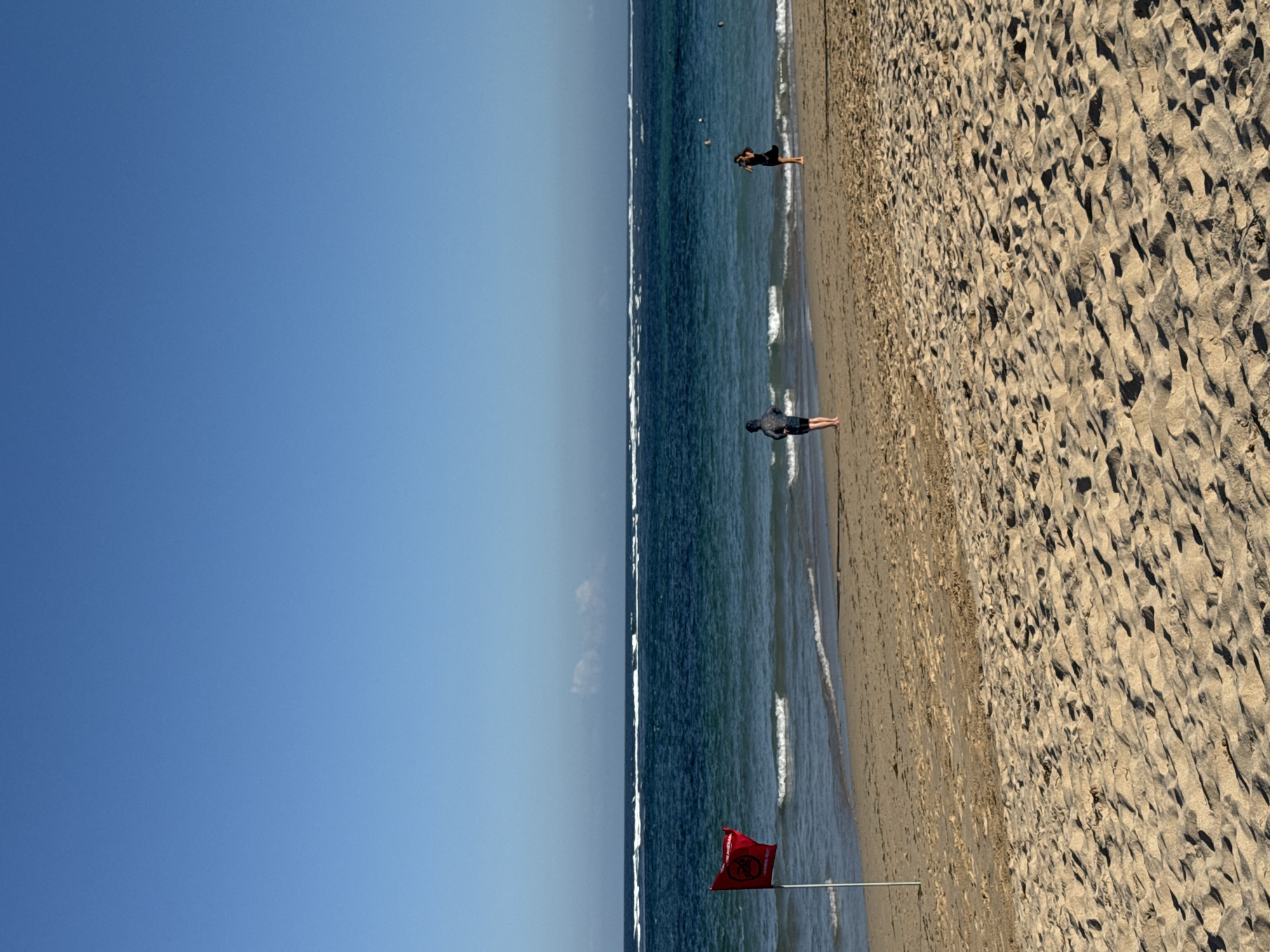 This is a photograph of a beach in Kuta Selatan, Indonesia. The sky is clear blue. The ocean is a deep blue with white waves. There is a red flag on a pole on the beach, indicating dangerous conditions. Two people are visible on the beach, and a few more are in the water. The sand is light brown and appears dry. The image has been cropped to focus on these elements.