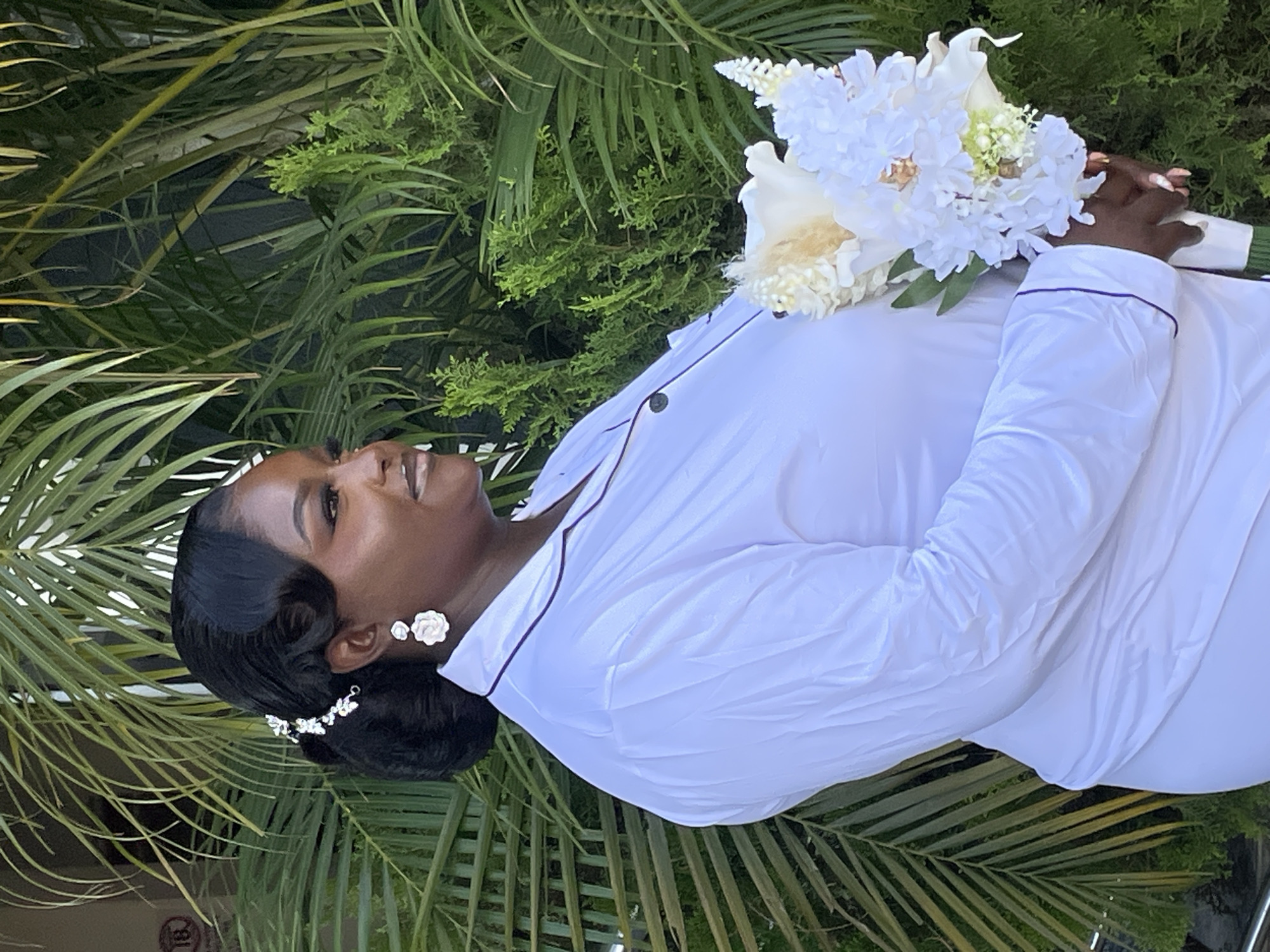 A woman holds a bouquet of white flowers, in what appears to be an outdoor area filled with leafy green plants, including palm fronds. She is wearing a white, long-sleeved shirt with dark piping and dark buttons. Her hair is styled in a dark updo and adorned with a silver hair clip. She is also wearing flower-shaped earrings. The location cannot be confirmed as Jos, Nigeria, as no other clues are present in the image.