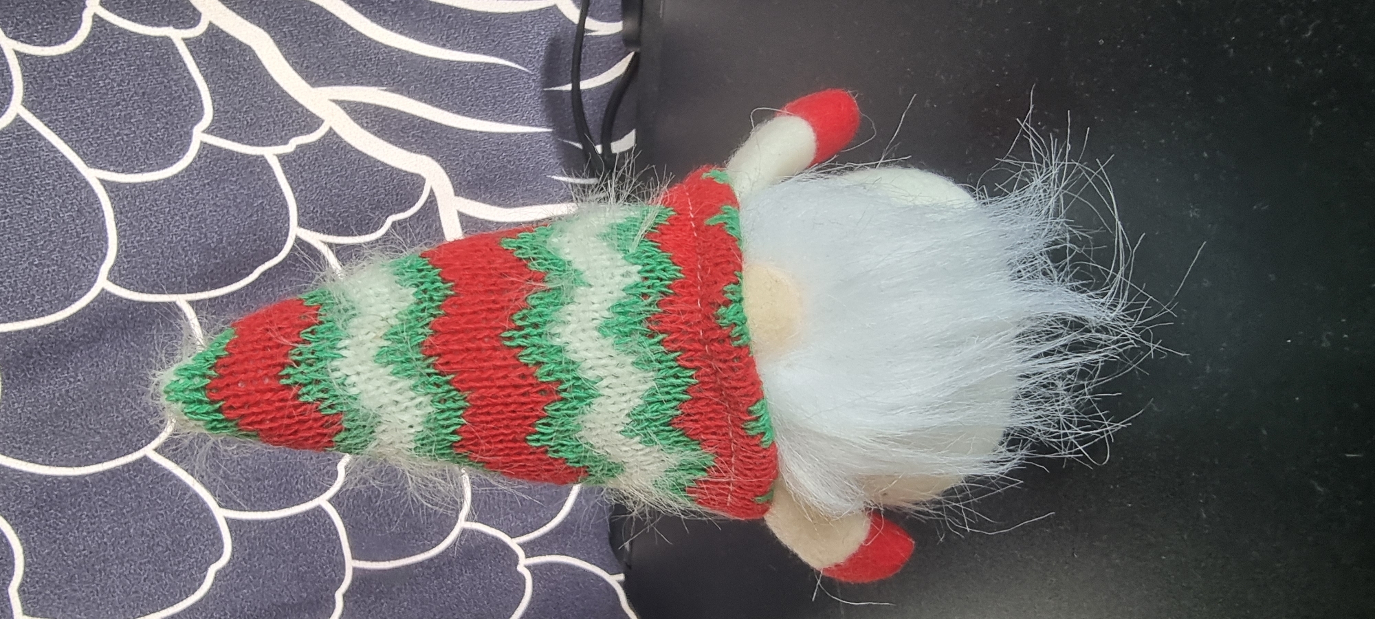 This is a close-up, horizontal shot of a gnome decoration. The gnome is positioned on its side, with its pointed hat extending towards the left of the frame and its fluffy white beard filling the right side. The hat is knitted in a zigzag pattern of red, green, and white stripes, and is trimmed with white fluffy material. The gnome has a small, round nose and red shoe-like feet. The background on the left is a dark blue fabric with a white abstract pattern. The background on the right is a dark, textured surface, possibly a table. The lighting suggests it is daytime, but there are no clear indicators of the time of day or weather. There is no visible text in the image. The setting appears to be indoors, possibly a home environment in Naaldwijk, Netherlands. The overall impression is that of a festive or decorative item.