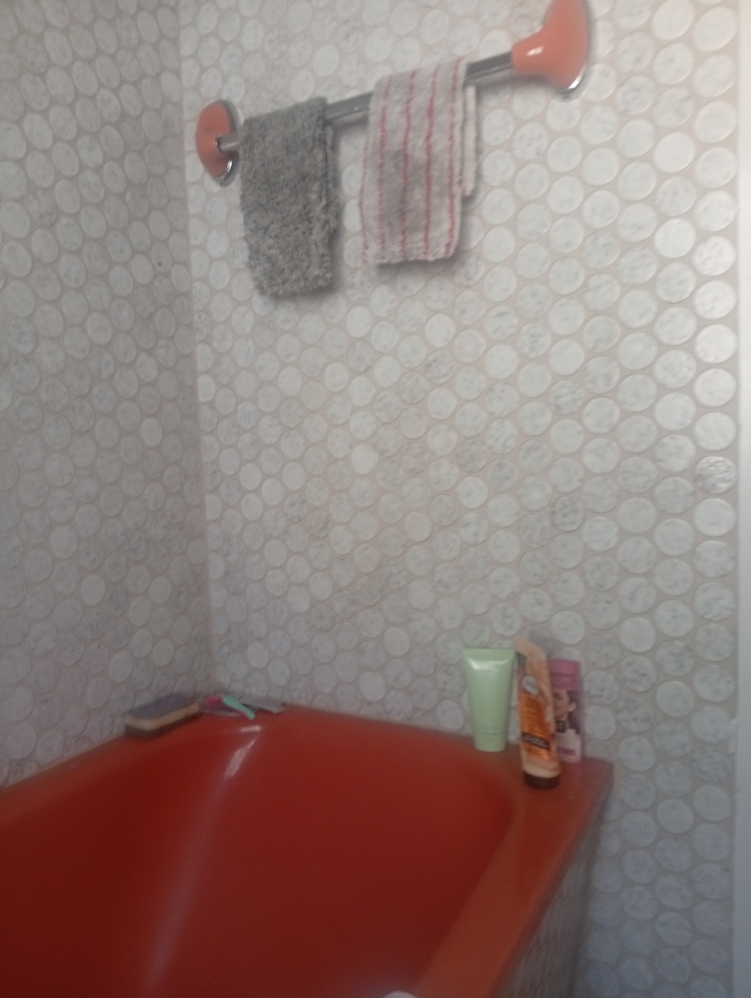 The image shows a close-up, eye-level view of a bathroom. In the foreground, a vibrant orange bathtub fills the bottom portion of the frame. Along the edge of the bathtub, on the right side, are several toiletries: a light green squeeze bottle, a tube of lotion with an orange label, and a small pink bottle. To the left of these, resting on the bathtub's edge, is a rectangular object that appears to be a bar of soap.
The background is dominated by tiled walls. The tiles are small, circular, and light-colored, arranged in a honeycomb pattern. Mounted on the tiled wall is a silver towel rack. Two towels are hanging on the rack. The towel on the left is dark grey and textured. The towel on the right is lighter with red stripes. Two orange towel holders are attached to the wall, one on each end of the rack.
There are no people visible in the image. The scene is static, suggesting a typical bathroom setting. The lighting is somewhat dim, casting reflections on the surface of the bathtub. No specific location in Kaiserslautern, Germany can be determined from the visual information provided.
