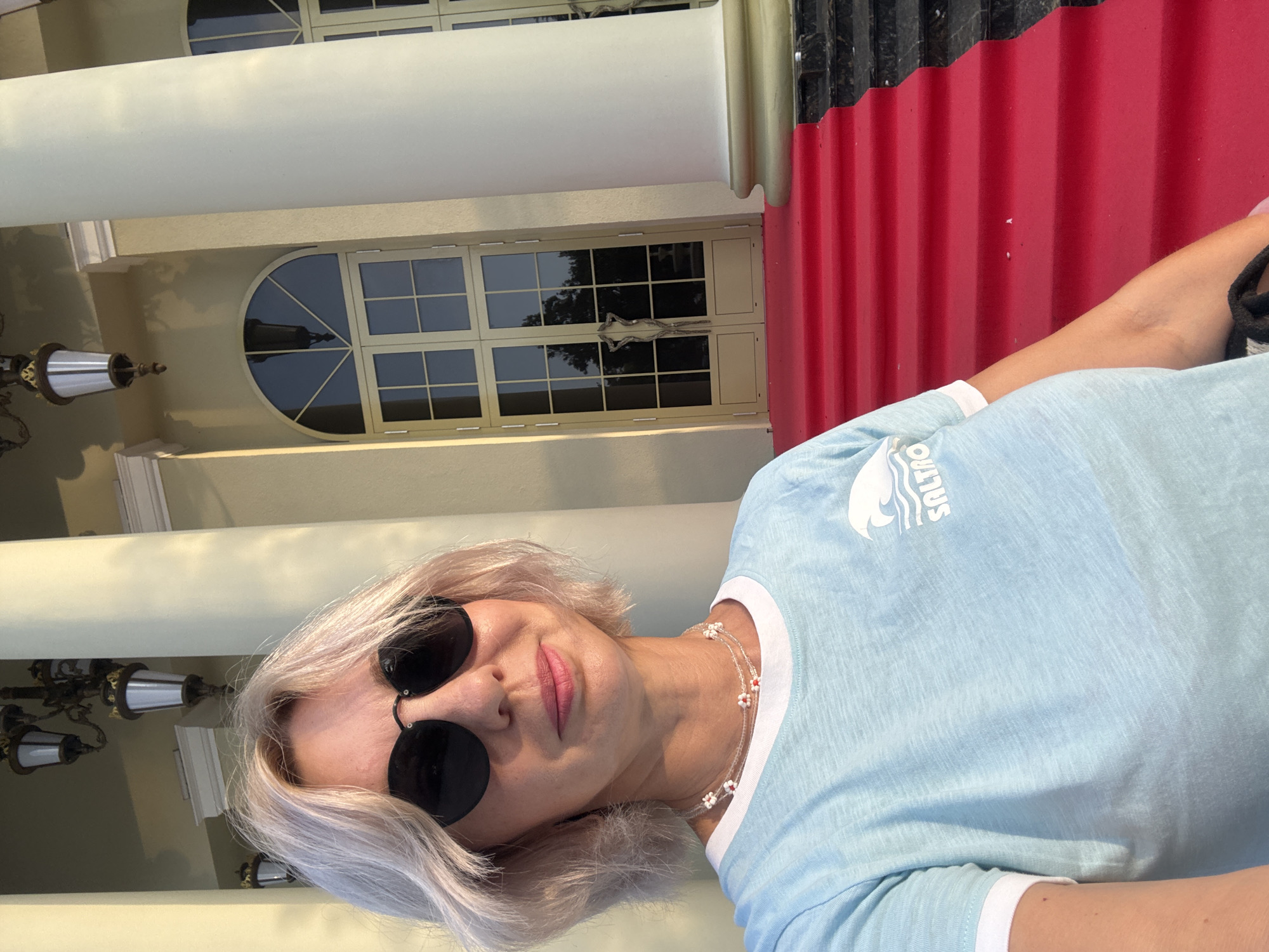 A woman with blonde, shoulder-length hair and sunglasses takes a selfie in front of a grand building with large columns and double doors. She is wearing a light blue t-shirt with a white logo and a beaded necklace. A red carpet leads up to the doors of the building, which appears to be in Sochi, Russia. The woman is smiling and looking at the camera. The lighting suggests it is daytime.