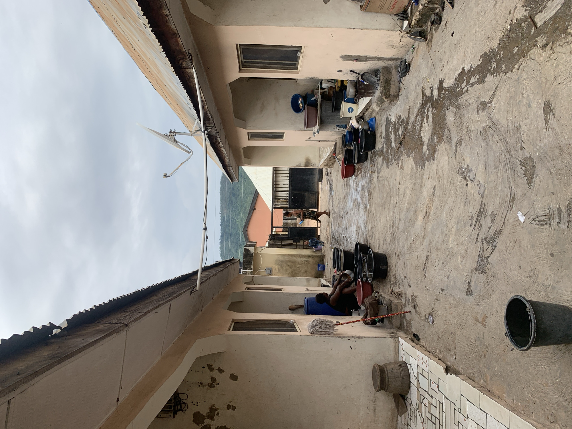 A daytime, tilted medium shot captures an outdoor courtyard in Lokoja, Nigeria, bordered by buildings with beige walls and corrugated metal roofs. A satellite dish is mounted on one of the roofs. The ground is concrete and wet, with visible streaks and patches. Several black buckets, along with a red one, are scattered along the courtyard. A man is seated, apparently washing in one of the buckets. Further down the courtyard, two other individuals are visible, one wearing patterned clothing. In the background, a gate is visible, beyond which lies a view of distant hills and buildings. The sky is overcast.
