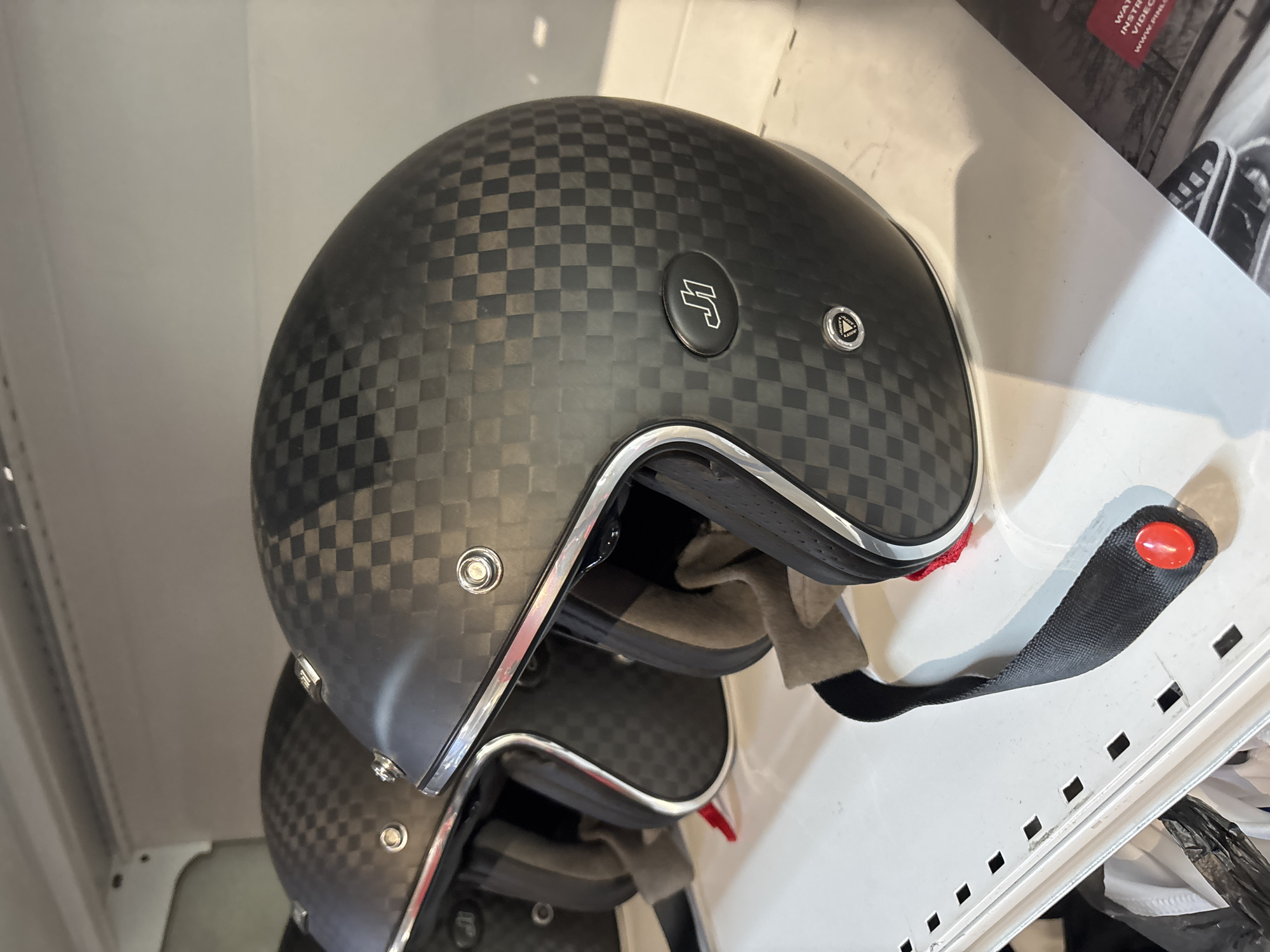 The image shows a close-up of a carbon fiber motorcycle helmet with a checkered pattern, displayed on a white shelf. The helmet has a chrome trim around the opening and a "J1" logo on the side. Another helmet is partially visible behind it. The setting appears to be a retail environment, possibly a store, given the shelving and the presentation of the product. There are no people or animals present in the image. The lighting is bright and even, suggesting an indoor setting. The exact location in Tbilisi, Georgia, cannot be determined from the visual information alone.
