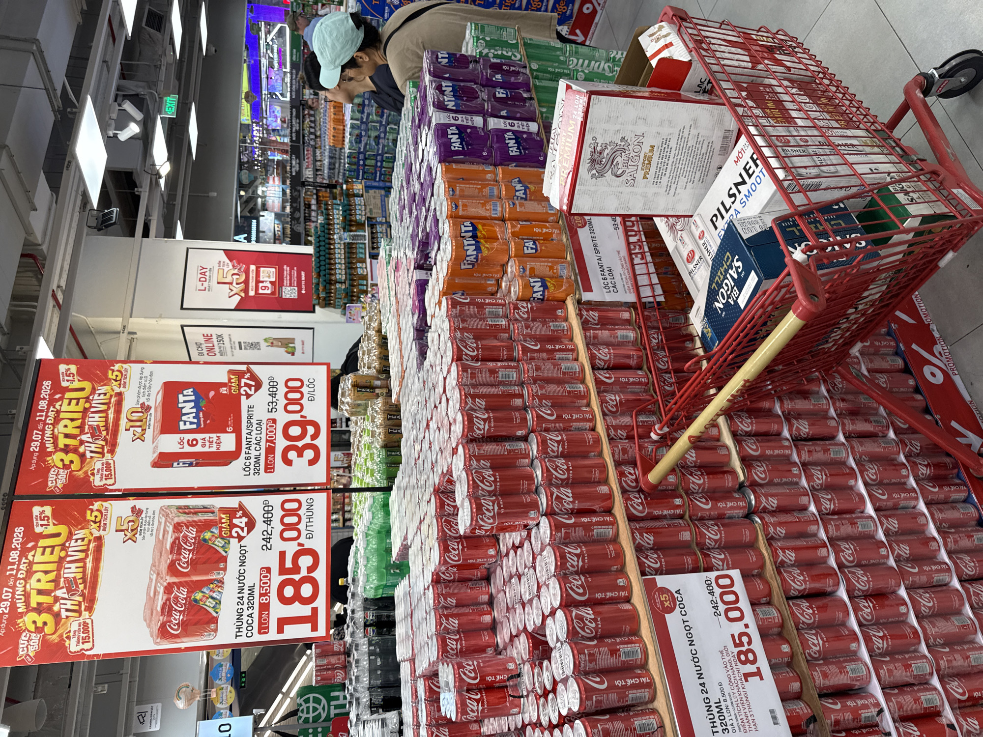 The image depicts a vibrant supermarket aisle in Nha Trang, Vietnam, showcasing a large display of Coca-Cola and Fanta beverages. Prominently featured are promotional signs advertising discounts and special offers on these drinks, including a "3 Triệu Thành Viên" (3 Million Member) campaign. The aisle is stocked with numerous cases and individual cans of Coca-Cola, Fanta, and Sprite, stacked high on shelves and in a red shopping cart. A few shoppers are visible in the background, browsing the products, one woman wearing a light blue hat. The overall environment is well-lit and organized, typical of a modern retail store.