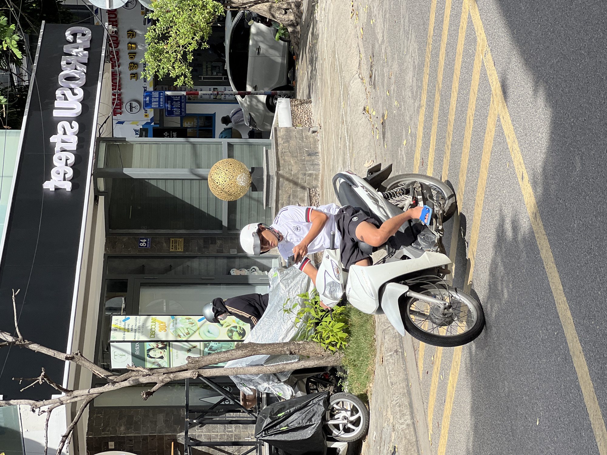 A young Vietnamese man, wearing a white t-shirt with red and black accents, black shorts, blue flip-flops, and a white helmet, rides a white motorcycle on a street in Nha Trang, Vietnam. He is looking down at his mobile phone, which he holds in his left hand. The motorcycle is white with black accents and has a dark seat. The street is paved with asphalt and marked with yellow lines. In the background, there is a building with a black facade and white lettering that reads "CAROS STREET". There is also a tree with bare branches on the left side of the image. To the right of the building, there is a white car parked on the street. Another person wearing a black hoodie and a silver helmet is partially visible on a motorcycle parked on the sidewalk. A black cover is on a motorcycle on the far left. The scene is illuminated by sunlight, casting shadows on the street.