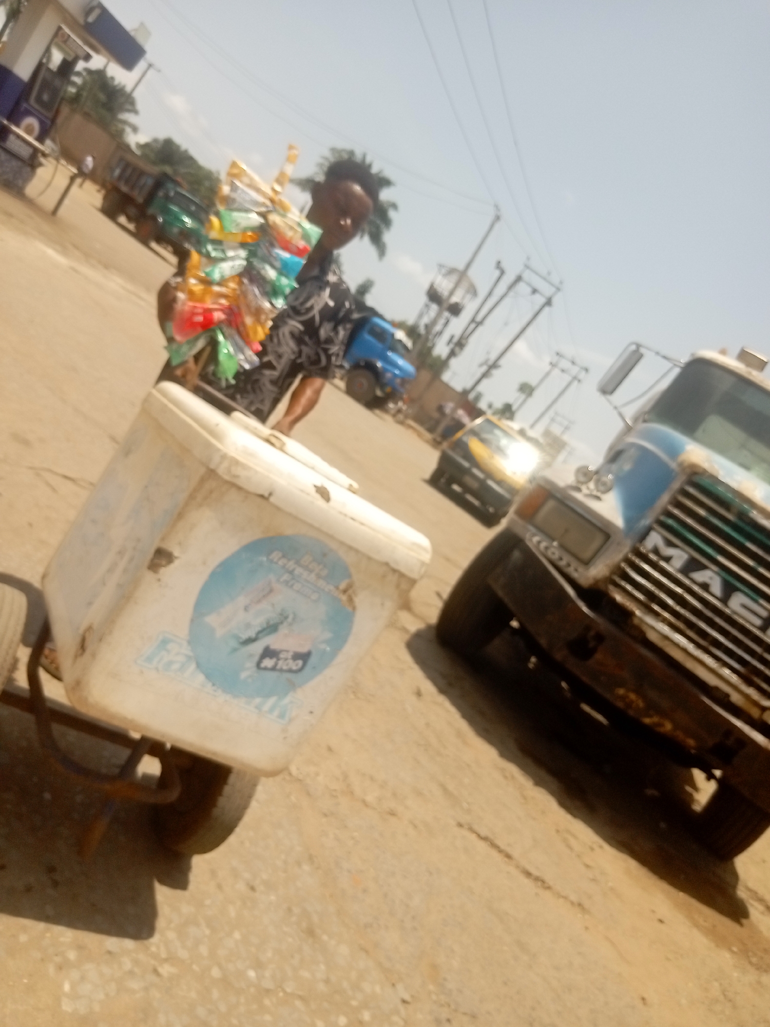 The image depicts a bustling street scene, characteristic of a commercial area, likely in Onitsha, Nigeria, under bright daylight. A street vendor is the central subject, positioned slightly left of center. The individual, dressed in a patterned dark top, carries a large, colorful display of various packaged snacks or candies on their person. In the foreground, a white cooler box on wheels, serving as a vendor's cart, is prominent. A blue circular logo on the cooler is visible, featuring text that includes "Refreshing" and "N100", with "Fast" written below it.
The background shows a dusty road with several vehicles. To the right, the front grille of a blue and white truck clearly displays the "MACK" logo. Further down the road, a yellow taxi is visible. On the far left, another large green truck and a structure resembling a gas station are in the distance. The sky is bright and clear, with utility poles and power lines running across the upper part of the frame. Strong shadows indicate a sunny day. The scene captures an everyday moment of street commerce.