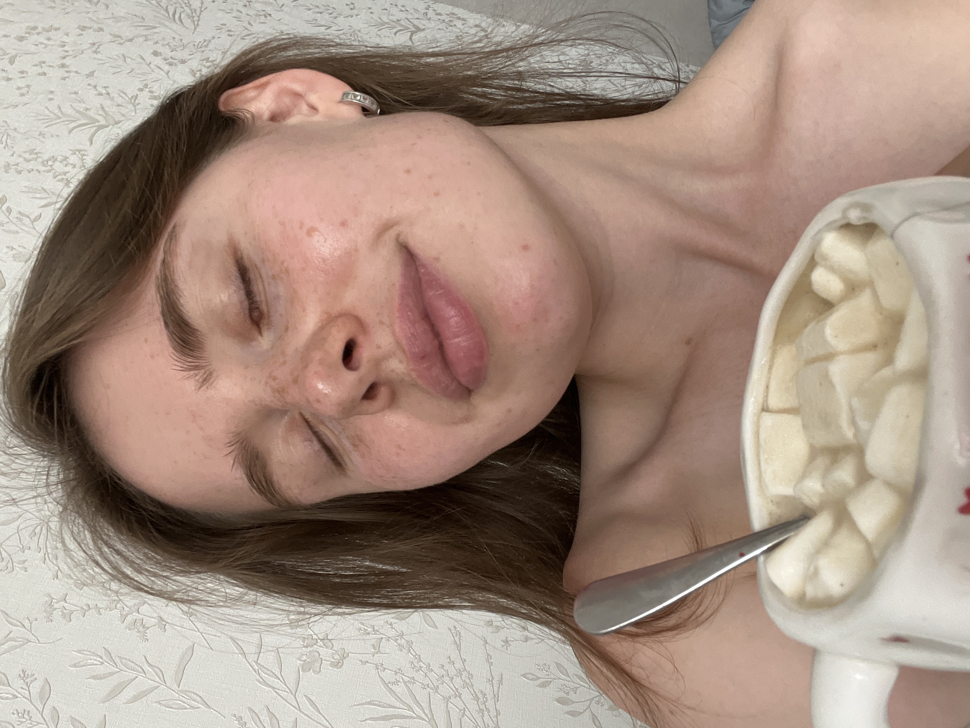 A young woman with brown hair and freckles is shown from the chest up, holding a mug of hot chocolate with marshmallows. She is looking down with her eyes closed and a slight smile. The background is a white wallpaper with a floral pattern. The image appears to be a selfie taken indoors.