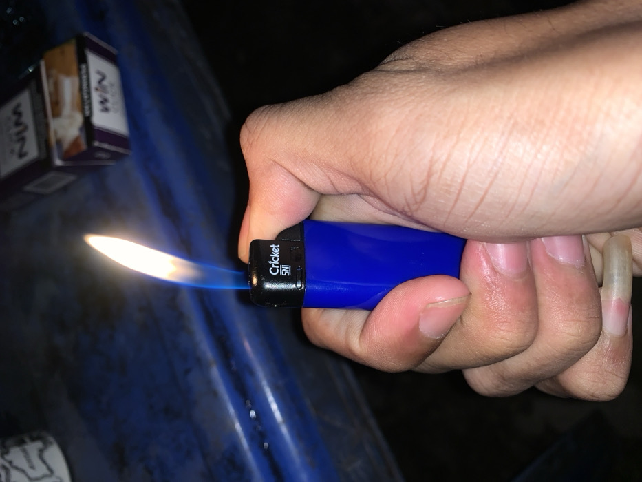 A close-up image shows a hand, likely an adult's, holding and operating a blue Cricket brand lighter, which emits a bright, elongated flame with a blue base and a yellowish-white tip. The lighter has the text "Cricket SNI" visible on its side, indicating compliance with Indonesian National Standards. The hand is positioned centrally in the frame, with fingers curled around the lighter. One fingernail, possibly on the pinky or ring finger, appears notably long and well-kept.
In the blurred background, two cigarette packs are visible, stacked or positioned next to each other, both bearing the brand name "WIN" in a prominent purple and white design. The setting appears to be dimly lit, suggesting either night time or an indoor environment with low ambient light, as evidenced by the vividness of the lighter's flame and the dark shadows. The surface beneath the lighter and cigarette packs is a dark blue, possibly textured material, resembling a table or plastic surface. The activity captured is the act of lighting the lighter, potentially in preparation to light a cigarette or for another purpose. The visible "SNI" mark and the cigarette brand "WIN" are consistent with the provided location context of Metro, Indonesia.