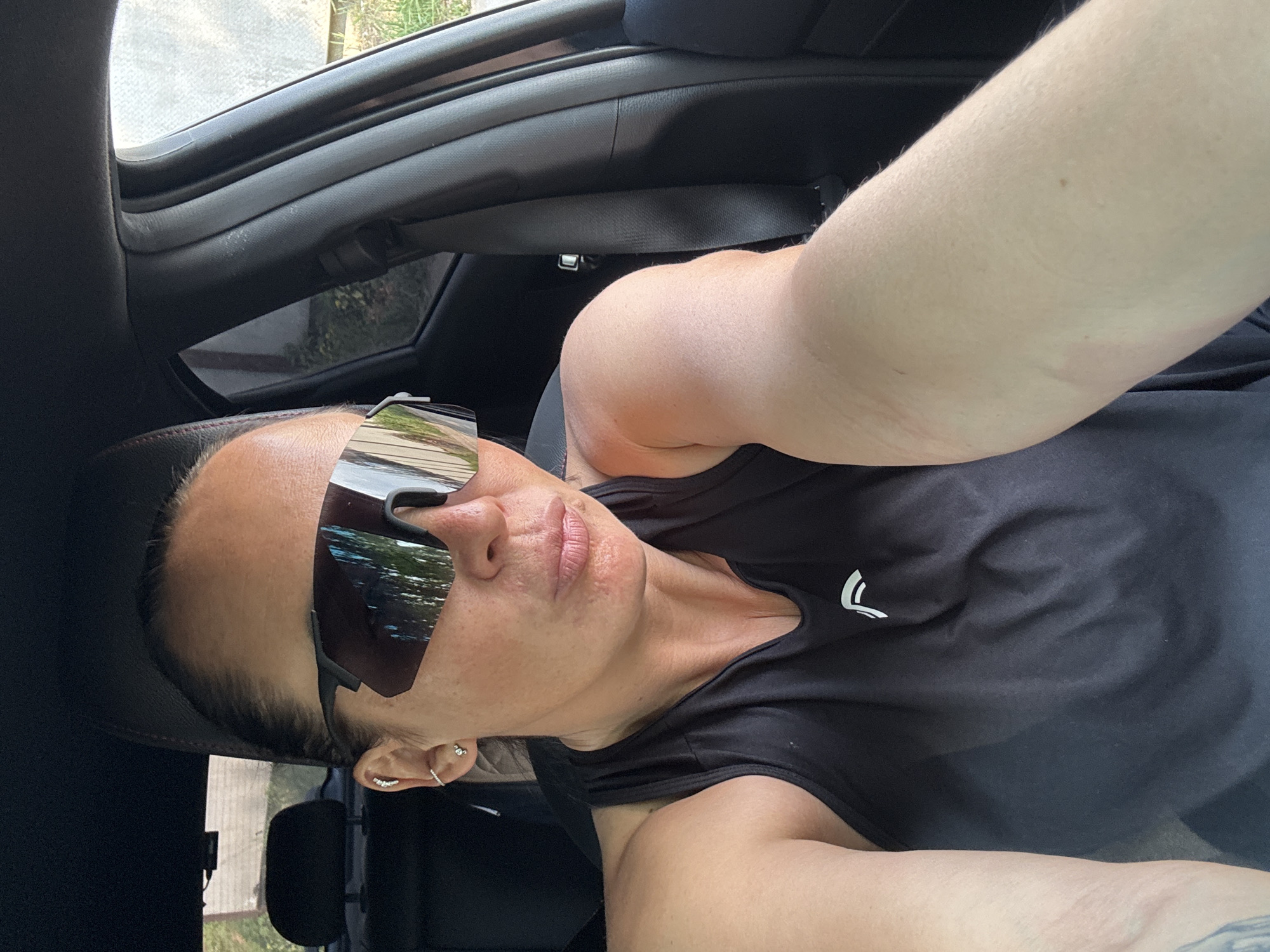 A selfie-style photograph shows a person with light skin wearing dark sunglasses and a black athletic shirt with a white logo on the chest, taken from inside a car. The interior of the car is dark, with the car door frame and roof visible. The reflection in the sunglasses suggests an outdoor setting with trees and possibly a street. The image does not provide enough information to determine the location as Chaikovsky, Russia.