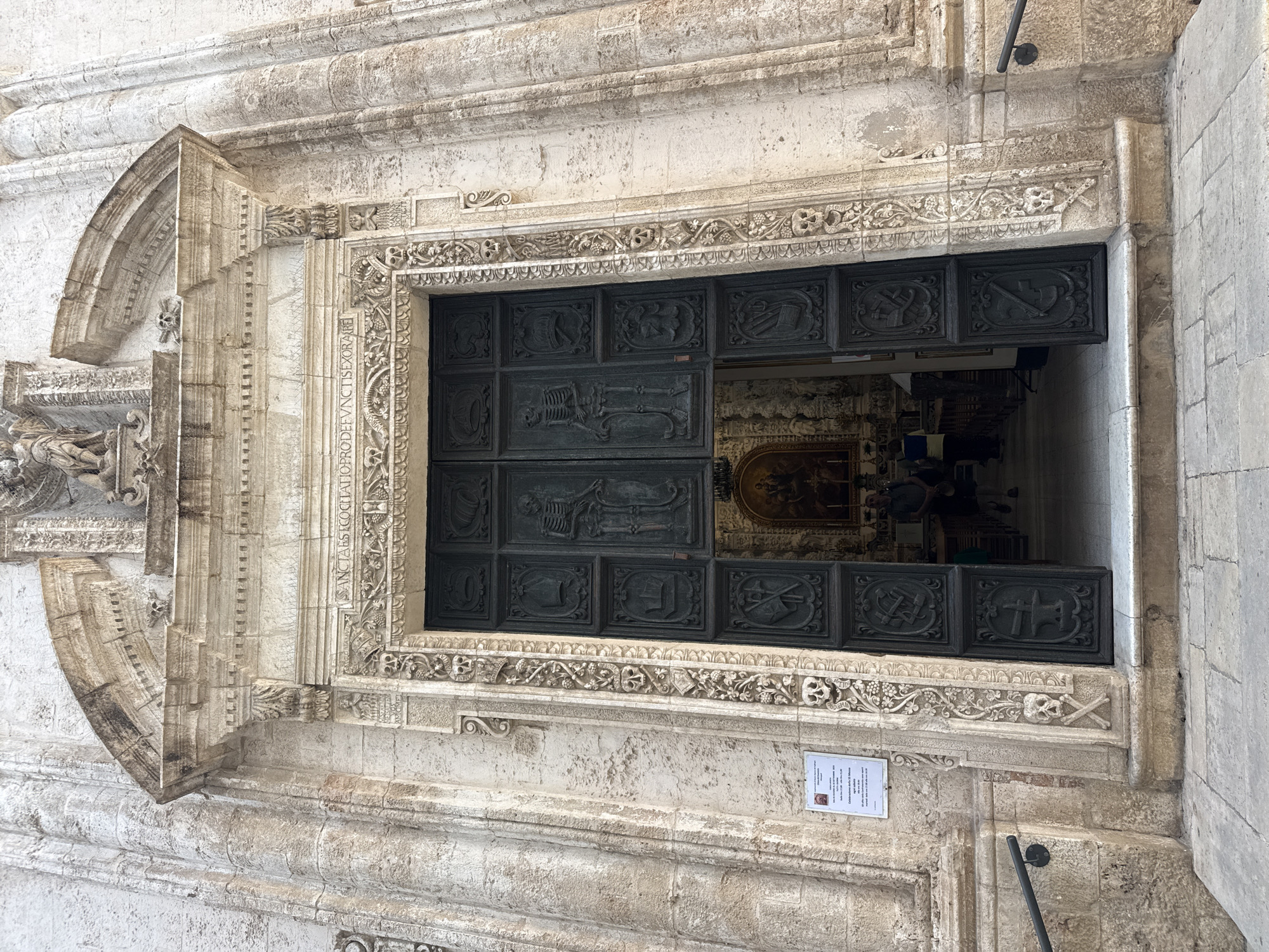 The image presents a grand, dark-colored door set into a weathered, light-stone facade, belonging to a historic religious building in Monopoli, Italy. The door, likely made of wood or metal, is composed of numerous rectangular panels, each featuring a carved relief. Notably, one panel prominently displays a full skeleton, while others depict skulls, crossbones, and tools such as a pickaxe, shovel, and scythe, serving as memento mori symbols.
The intricate stone facade surrounding the door is adorned with elaborate floral motifs, scrollwork, and a frieze above the entrance that also incorporates skull and crossbone imagery. A Latin inscription, partially visible on the left vertical frame of the door, reads "QATIO.PRO.DEFVNC.TIS.EXORA.R," suggesting prayers for the deceased.
The door is partially open, revealing a glimpse into the opulent interior of what appears to be a church or cathedral. A large, gilded altarpiece or artwork is visible deep inside, and several indistinct figures of people can be seen, indicating ongoing activity within the building.
Below the door, a small white printed sign is affixed to the stone wall. The word "MONOPOLI" is discernible at the top, along with "Cattedrale della... Monopoli," confirming the location and type of structure. The sign also features a small photograph of a person and Italian text, likely an announcement or schedule. The scene is illuminated by natural daylight, suggesting a clear day. The overall impression is one of historical grandeur and solemnity.