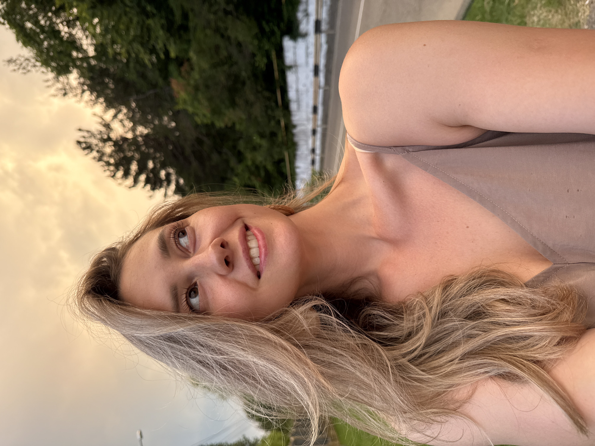 A young woman with blonde hair and fair skin is photographed outdoors during the golden hour. She is wearing a light brown tank top and is looking upwards and to her right with a slight smile. Her hair is windswept and falls over her shoulder. In the background, there is a road bordered by a low white wall and railings, with a dense line of trees beyond that. The sky is cloudy and illuminated by the setting sun, casting a warm glow over the scene. The location is the city of Nebug, Russia.