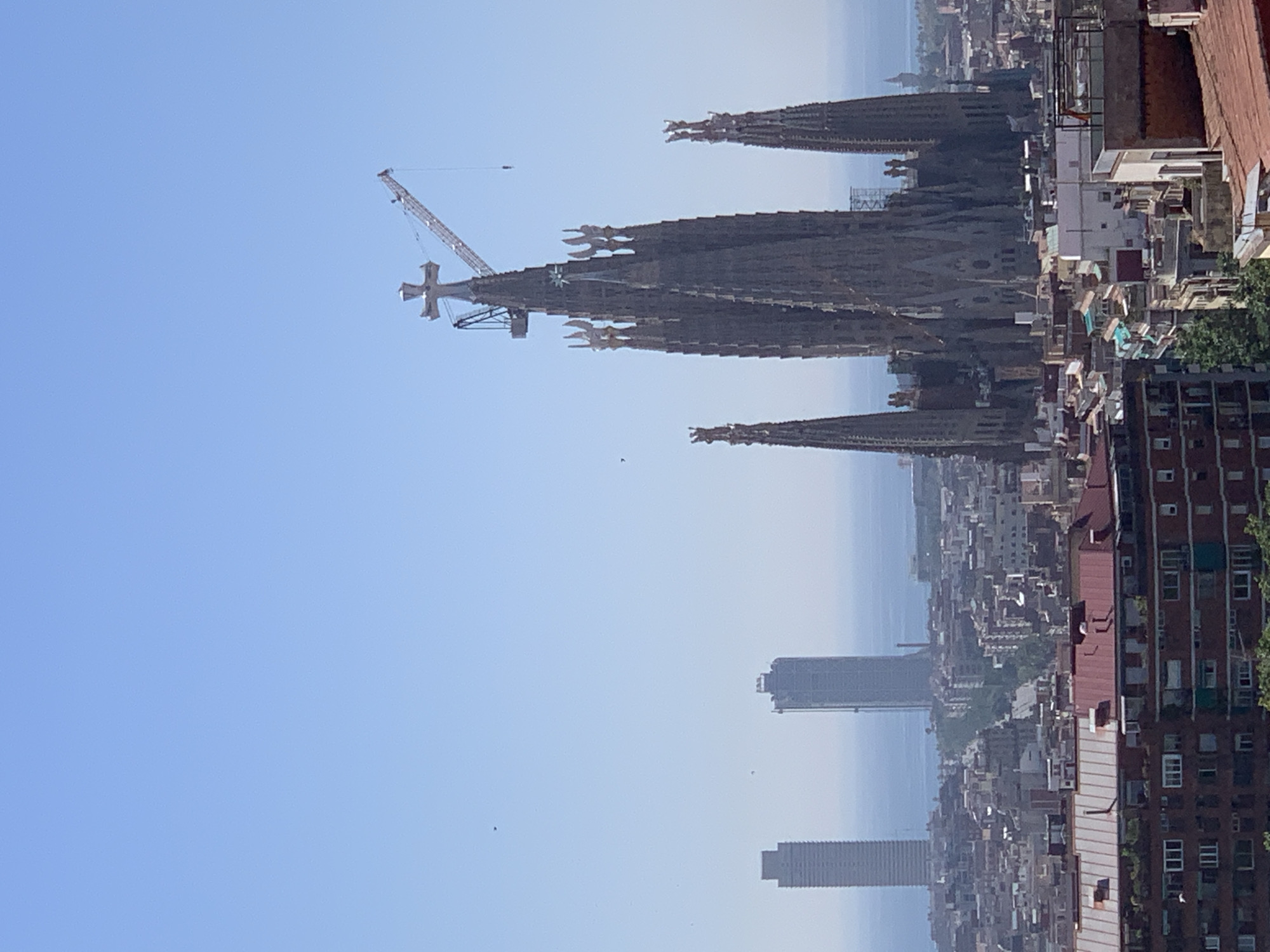 This is a daytime, aerial view of the Sagrada Familia basilica in Barcelona, Spain, under construction, as indicated by the presence of a large crane on top of one of its towers. The basilica's distinctive spires dominate the center of the image, with its intricate Gothic architecture visible. In the background, two modern skyscrapers are visible on the left, along with the blue waters of the Mediterranean Sea. The foreground and middle ground are filled with a dense urban landscape of residential buildings with red-tiled roofs, typical of Barcelona. The sky is clear and blue. There are no people or animals clearly visible in the image.