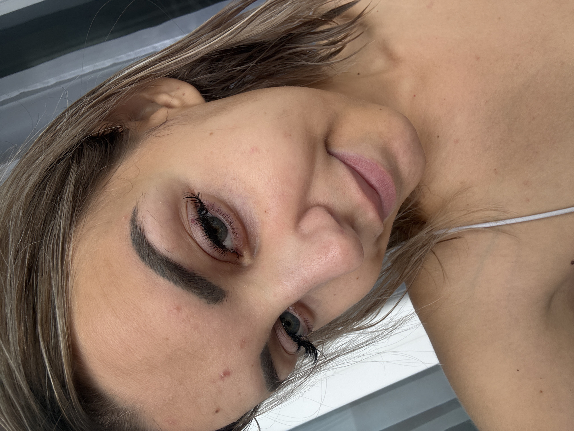 The image is a close-up selfie of a young woman with brown hair and fair skin. She has a slight smile and is looking down and to the left. Her eyes are open, and she is wearing dramatic false eyelashes. There are a few small blemishes on her forehead and cheek. She is wearing a white tank top. The background is out of focus and appears to be a striped curtain or blinds. The location in Kholmsk, Russia is not discernible from the image.