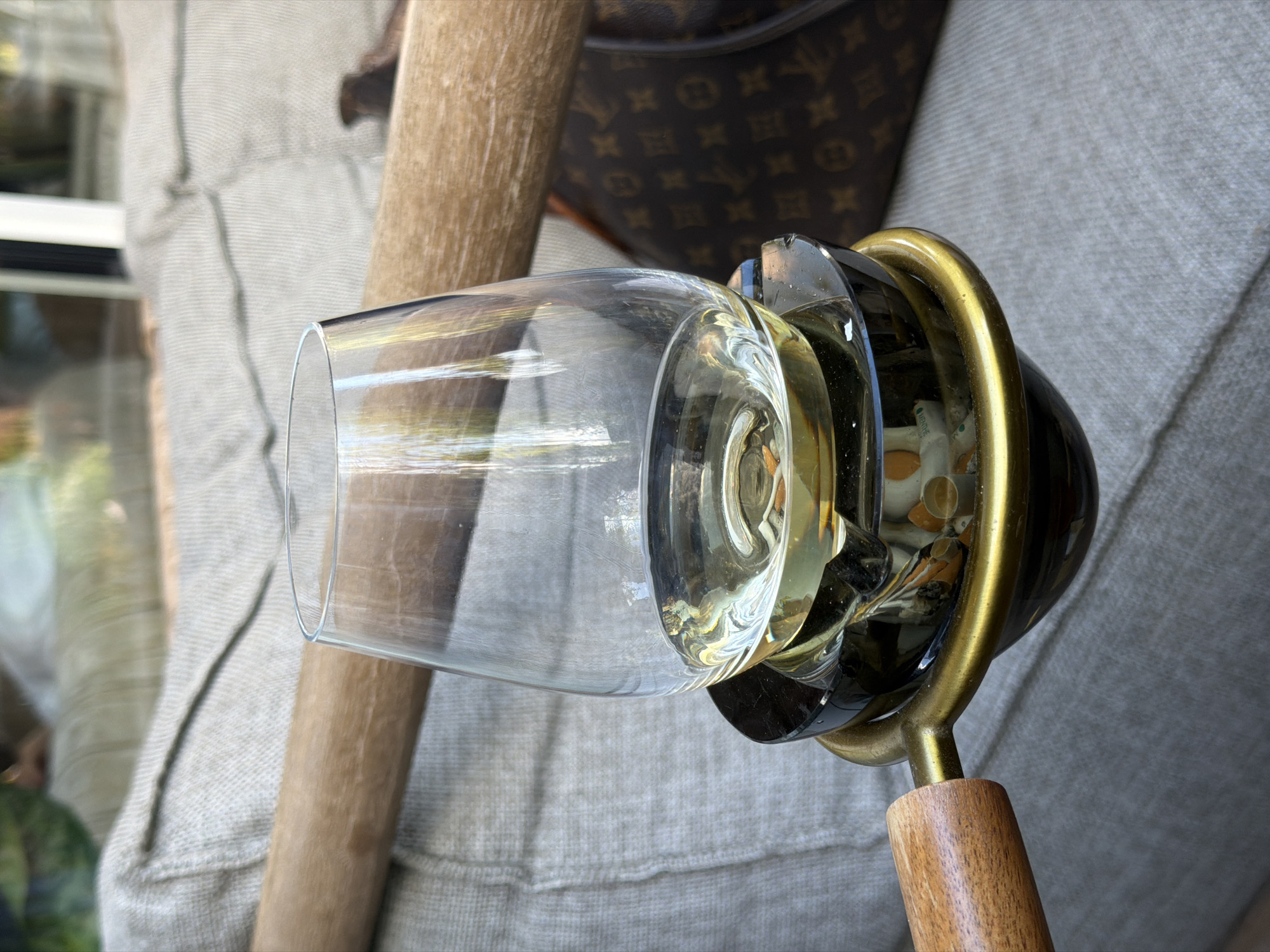 A still life of a glass of white wine tilted slightly, within a brass and black metal frame, with a wooden handle. The glass contains a light-colored liquid and the frame appears to be a holder or stand for the glass. In the background, out of focus, are elements including a wooden post, a brown patterned bag (possibly a designer handbag), and grey fabric that looks like upholstery. The setting is indoors, with soft, natural lighting. The image appears to be close-up, focusing on the wine glass and its holder. It is not possible to determine from the image if it is located in the city of London, United Kingdom.