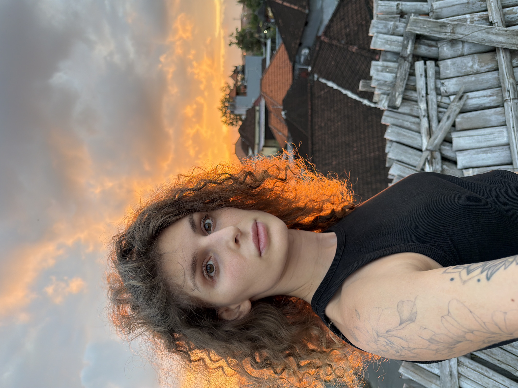 A medium shot captures a woman with curly brown hair, wearing a black tank top, against a sunset sky filled with orange and gray clouds. Her face is turned towards the camera, with her gaze directed forward. Her left arm is visible, displaying a tattoo of floral outlines. In the background, several rooftops with terracotta tiles are visible, suggesting an urban or suburban setting. A weathered wooden fence made of bamboo poles is positioned in the foreground, partially obscuring the view of the rooftops. The lighting indicates it is either sunrise or sunset. The location is identified as North Kuta, Indonesia.