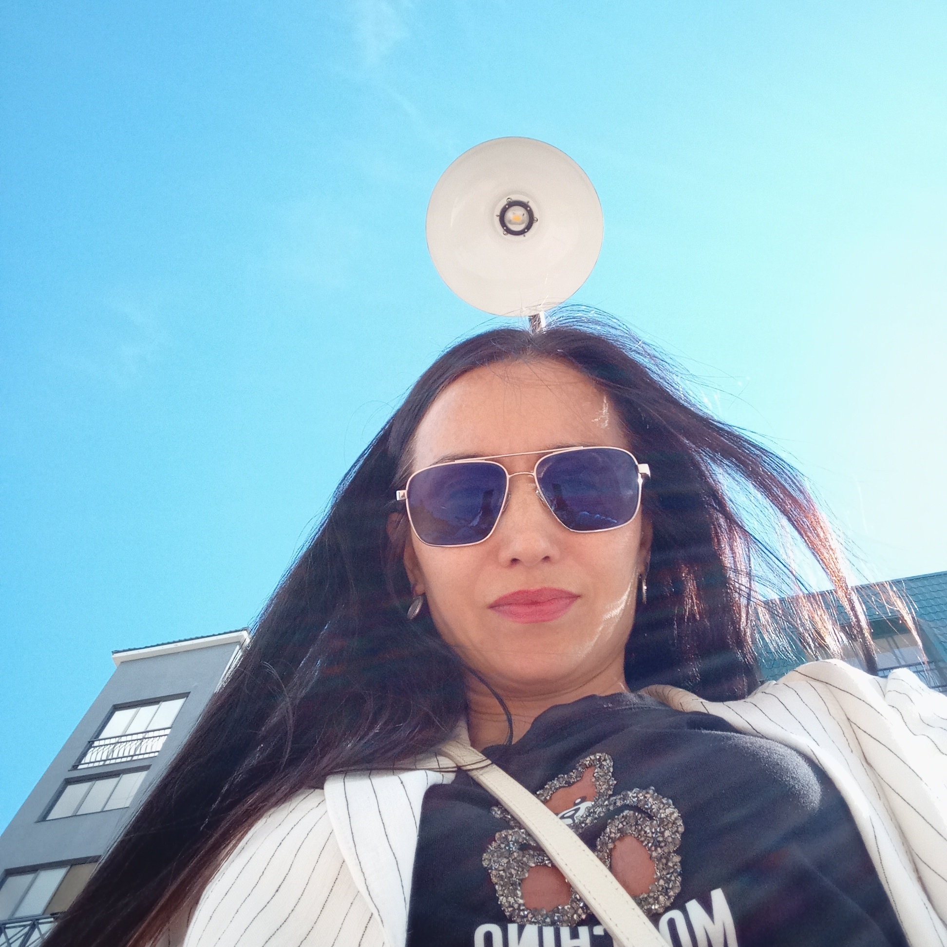 A low-angle portrait captures a woman outdoors in Pervomaysky, Russia, under a bright, clear blue sky with minimal cloud cover. The woman, with long, dark hair flowing around her face and shoulders, looks directly at the camera. She wears large, reflective blue aviator-style sunglasses with a metallic frame, obscuring her eyes. Her lips are painted with a reddish-pink lipstick, and she has small earrings.
She is dressed in a black top, partially visible, which features a prominent, glittery or rhinestone-embellished design. Below the design, upside-down letters "OHIW" are visible, likely part of a brand name. Over her top, she wears a light-colored, possibly white, blazer or jacket with subtle vertical stripes. A light-colored strap, likely from a shoulder bag, crosses her chest.
Directly above her head, positioned against the expanse of the sky, is a circular white lamp or light fixture with a visible central LED bulb. In the background, to the left, a modern grey building with white-framed windows and a small balcony with a black railing is visible, suggesting an urban or suburban setting. The bright sunlight indicates it is daytime, likely midday or early afternoon, creating clear contrasts and highlights. The woman's expression is neutral, partially masked by her sunglasses. The overall impression is a casual outdoor portrait on a sunny day.