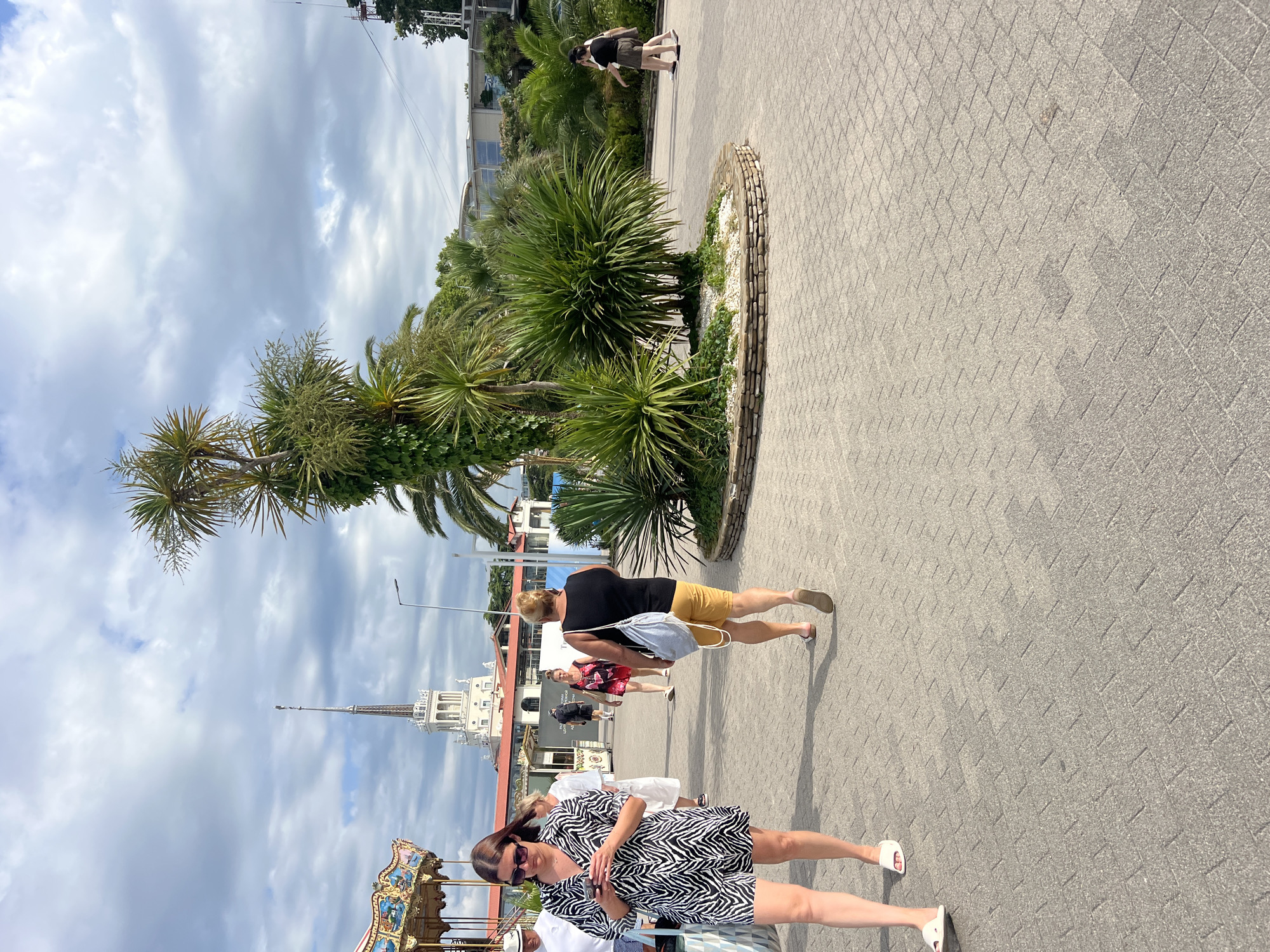 The image depicts a scene in Sochi, Russia, likely near the port area, with a prominent white tower featuring a spire in the background. A carousel with colorful decorations is visible to the left. The foreground is a paved walkway with several people strolling. A woman in a zebra-print dress is looking at her phone, while another woman in a black tank top and yellow shorts walks away from the camera, carrying a light blue bag. Further in the background, more people are walking, and palm trees and lush green foliage are interspersed with buildings. The sky is cloudy, suggesting a dynamic weather day. The overall atmosphere appears to be a public outdoor space, possibly a promenade or a park area within the city.