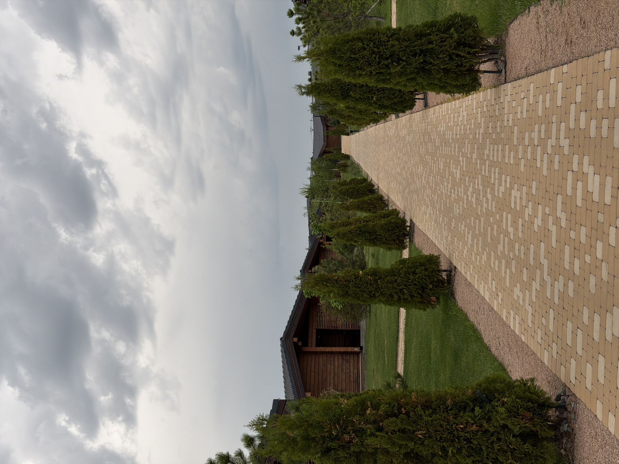 The image shows a path made of tan and cream pavers leading towards a wooden structure with a dark roof, likely a cabin or lodge, in what appears to be a landscaped garden. The path is flanked by rows of small, conically shaped evergreen trees, and the green lawn is visible on both sides. The sky is overcast with gray clouds, suggesting a cloudy or possibly stormy day. There are no people visible in the image. The exact location within Kamyshin, Russia cannot be determined from the given image.
