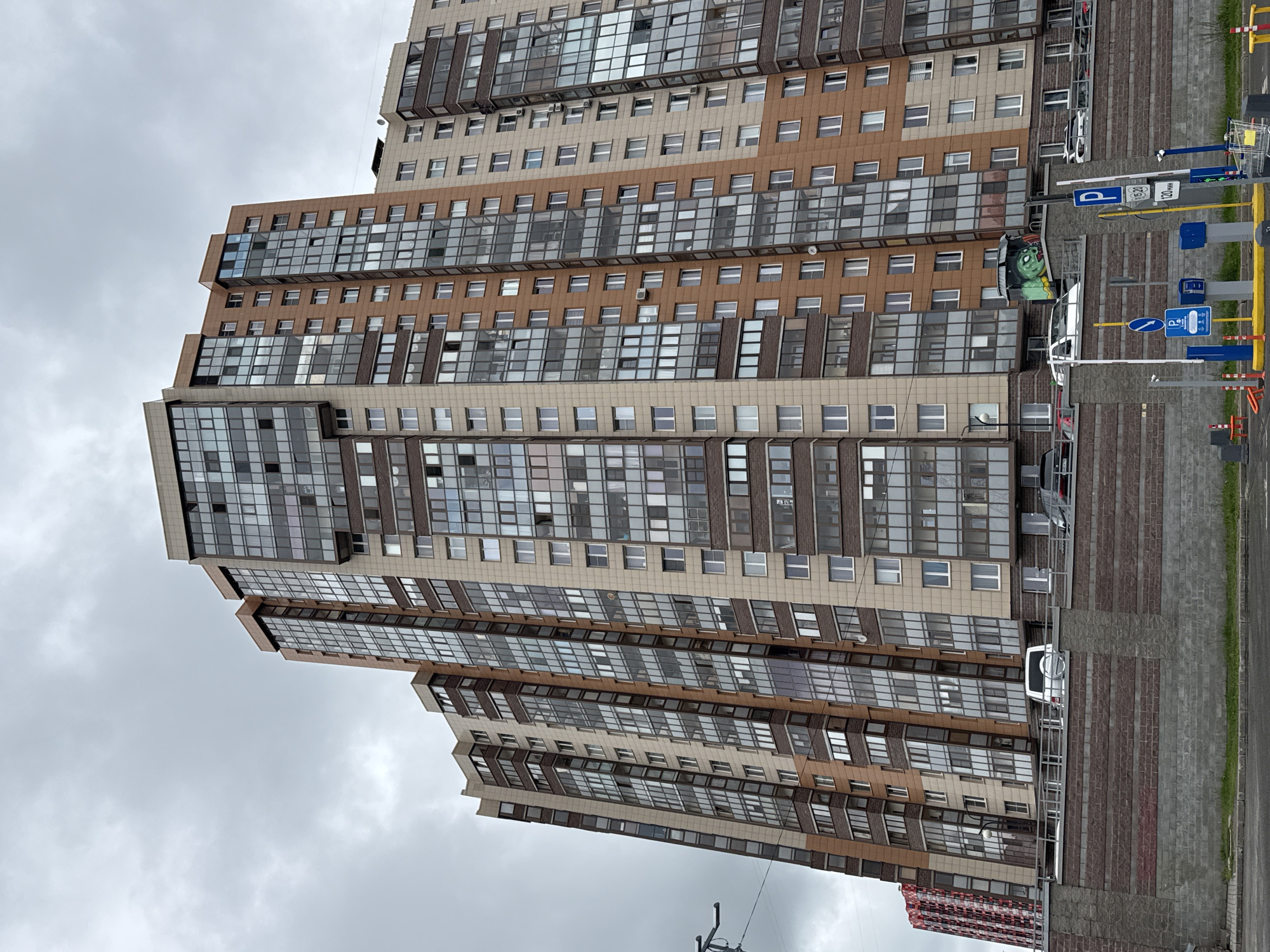 The image shows a high-rise apartment building in Saint Petersburg, Russia. The building has a stepped design, with alternating floors of brown and beige-colored panels, interspersed with rows of glass windows. There are several parked cars adjacent to the building. The sky is overcast. A parking sign and other directional signs are visible near the building. A partial view of another, smaller building with a distinctive red and white pattern can be seen in the bottom right corner.
