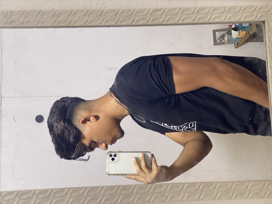 An indoor mirror selfie captures a young adult male in profile, facing left, within a residence in Juan Griego, Venezuela. He is holding a light-colored smartphone with a clear case in his extended right hand, aiming it at his reflection. The male has dark, wavy hair with a prominent fade haircut and a subtle goatee. He wears a black short-sleeved t-shirt with a partial white graphic design on the left chest and a silver-colored chain necklace. The background reflected in the mirror is a plain, light-colored wall featuring a dark circular mark in the upper central area. The mirror itself has a light-colored, decorative frame with a repetitive wavy pattern. To the lower right in the reflection, a small table or shelf displays a framed picture and several small containers.