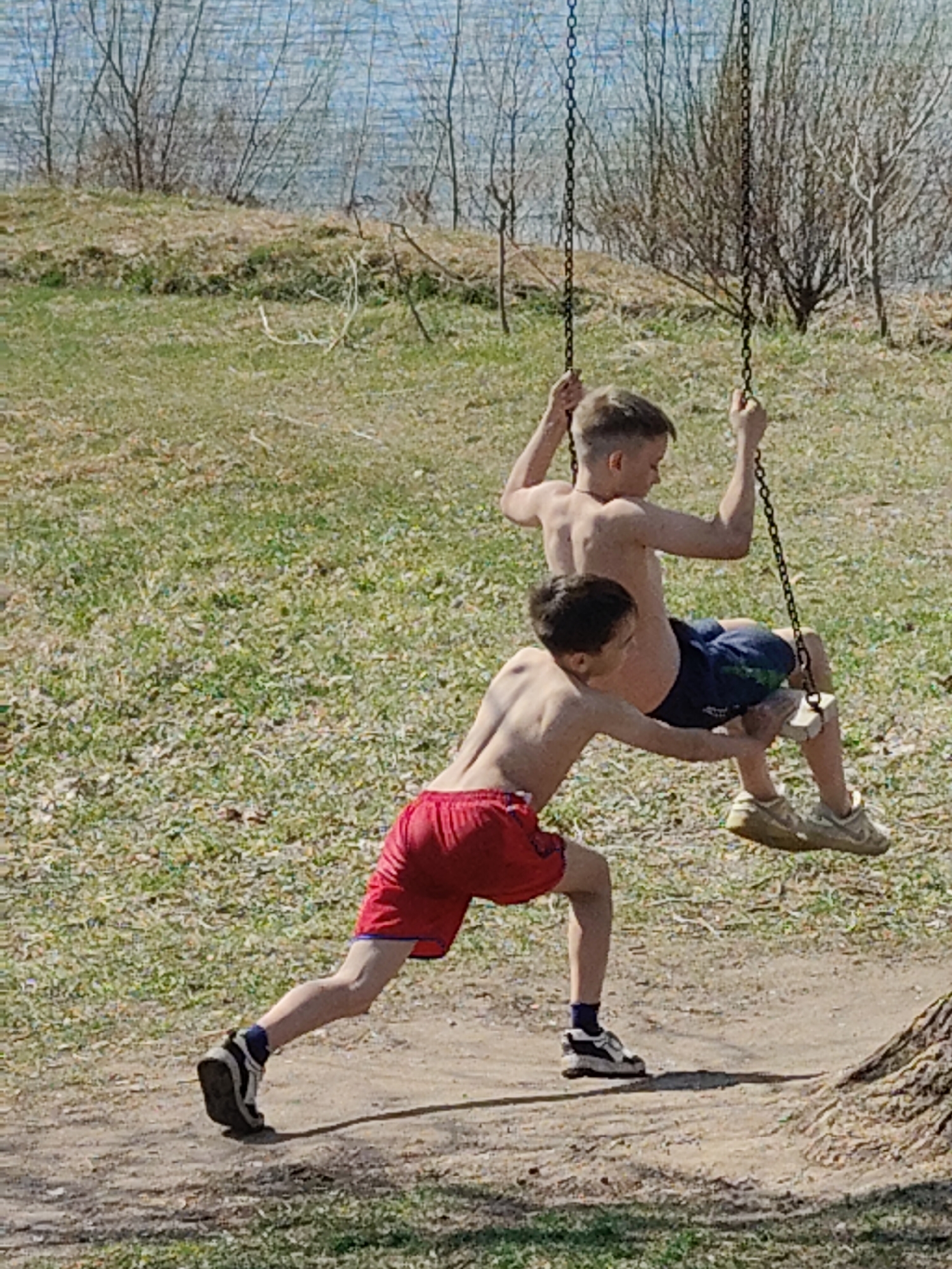 Two shirtless boys are on or near a swing set. One boy is seated on the swing, wearing blue shorts and tan sneakers. The other boy stands behind him, wearing red shorts, black socks, and black and white sneakers, and appears to be pushing the swing. The location appears to be an outdoor area with grass, dirt, and some trees, possibly near a body of water.