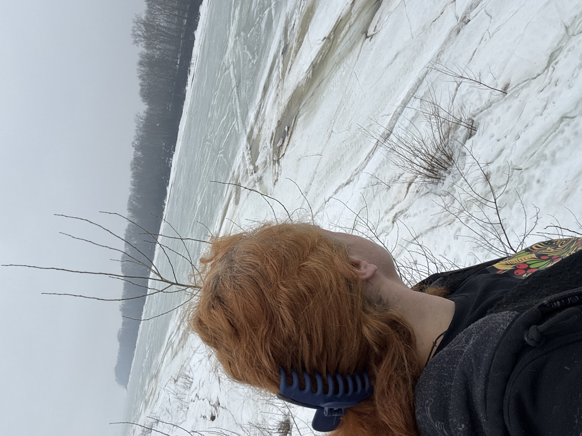 A person with reddish-brown hair, secured by a dark blue hair clip, is positioned in the foreground against a snow-covered landscape. The person is wearing dark clothing and appears to be lying down. In the background, there is a partially frozen body of water with cracked ice surfaces. A line of trees is visible along the distant bank of the water. The scene suggests a cold, wintry environment. Based on the context, it appears this photo was taken in Chaikovsky, Russia.