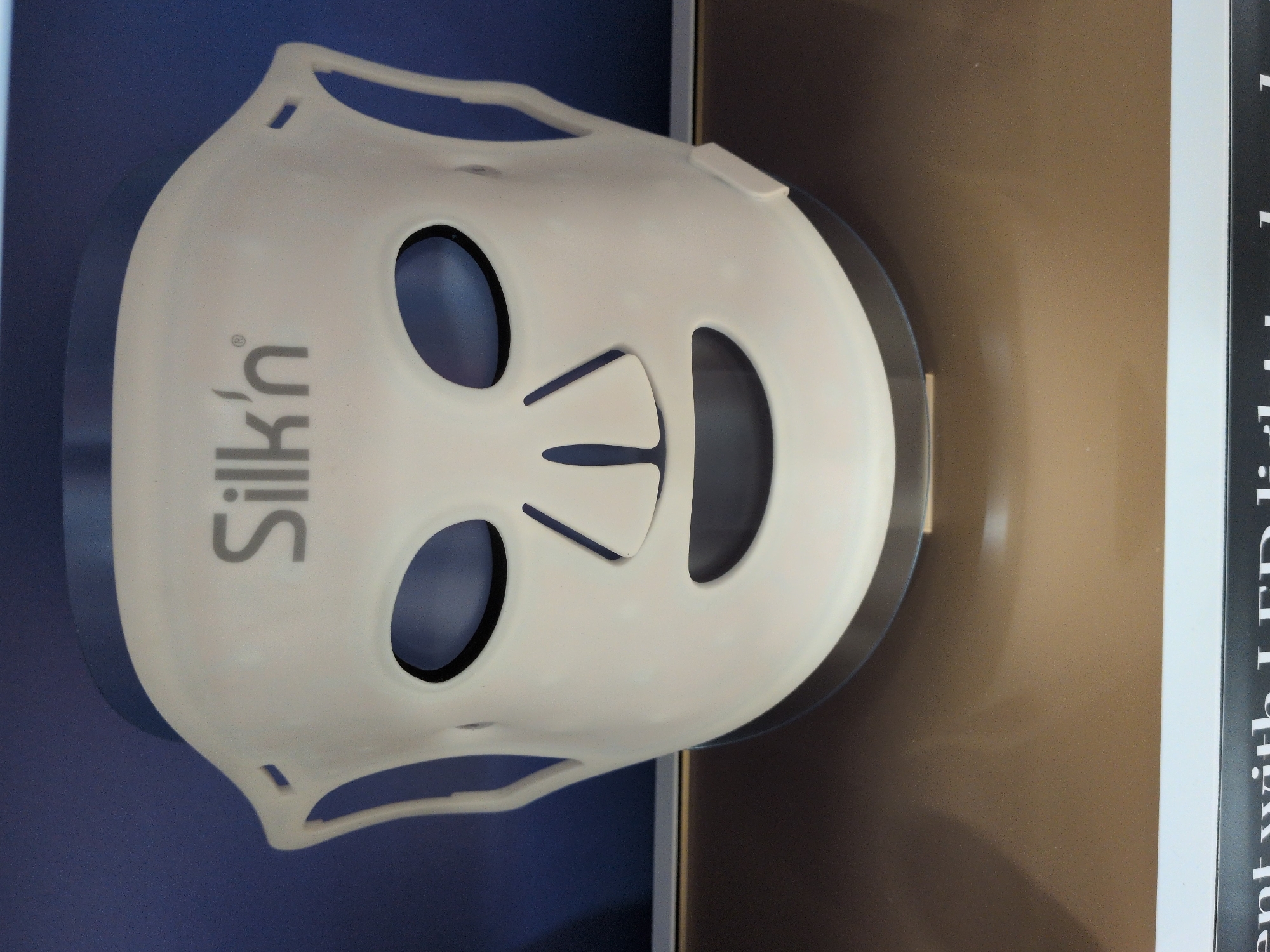 A white Silk'n face mask with eye and mouth openings is displayed against a dark blue background and a reflective brown surface. The mask has the brand name "Silk'n" printed in grey letters.