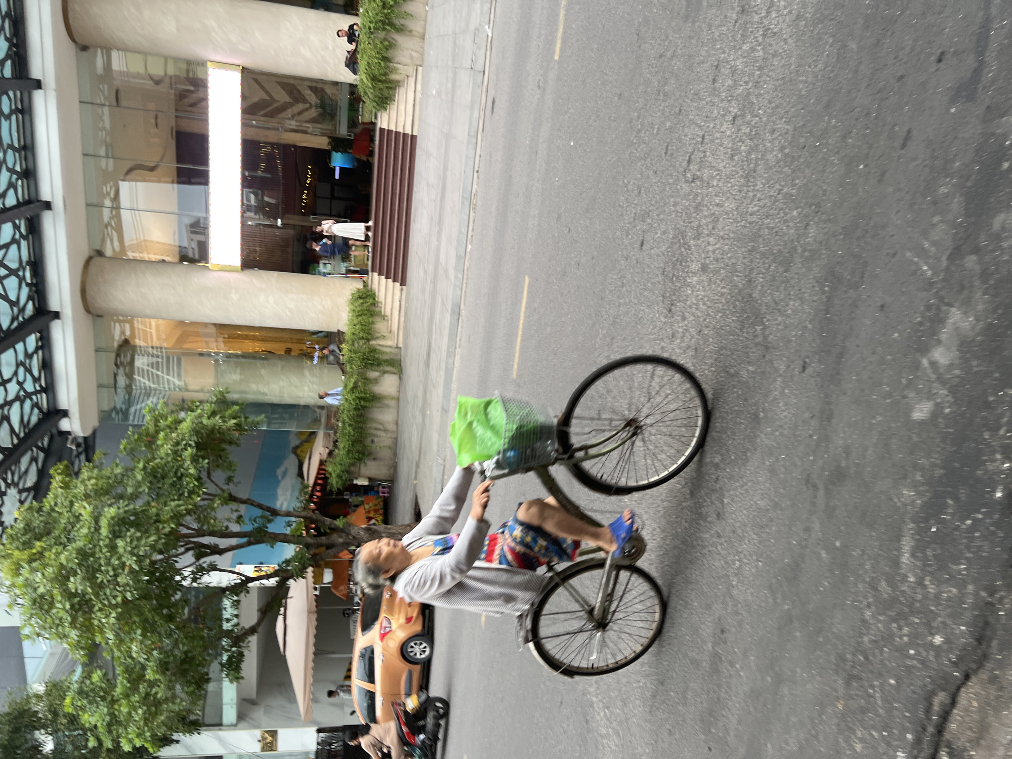 In Nha Trang, Vietnam, an elderly woman cycles down a paved street on a bicycle. She is wearing a grey cardigan, a colorful patterned dress, and blue flip-flops. A green bag is attached to the front of her bicycle. The street is bordered by a sidewalk, lush green plants, and a modern building with large columns and glass windows. A light-colored car is parked on the street to the left of the woman. The sky is overcast, suggesting a cloudy day.